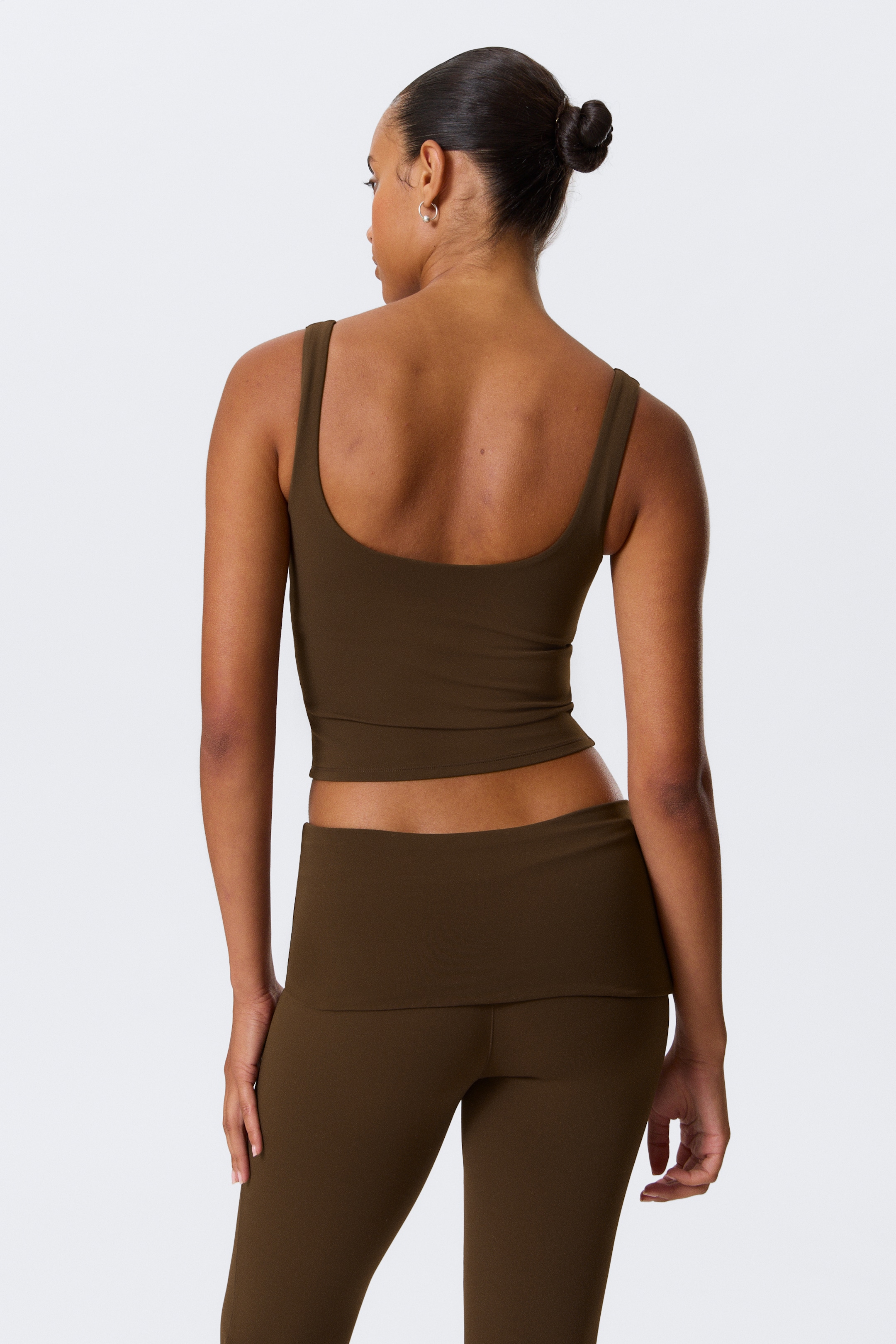 Grotere afbeelding bekijken: Een bijpassende set van een donker olijfkleurige tanktop en legging wordt gepresenteerd vanuit een achteraanzicht. De tanktop heeft brede schouderbanden en een ondiepe ronde rug, eindigend op een kortere lengte, terwijl de legging een brede, omvouwbare tailleband toont.