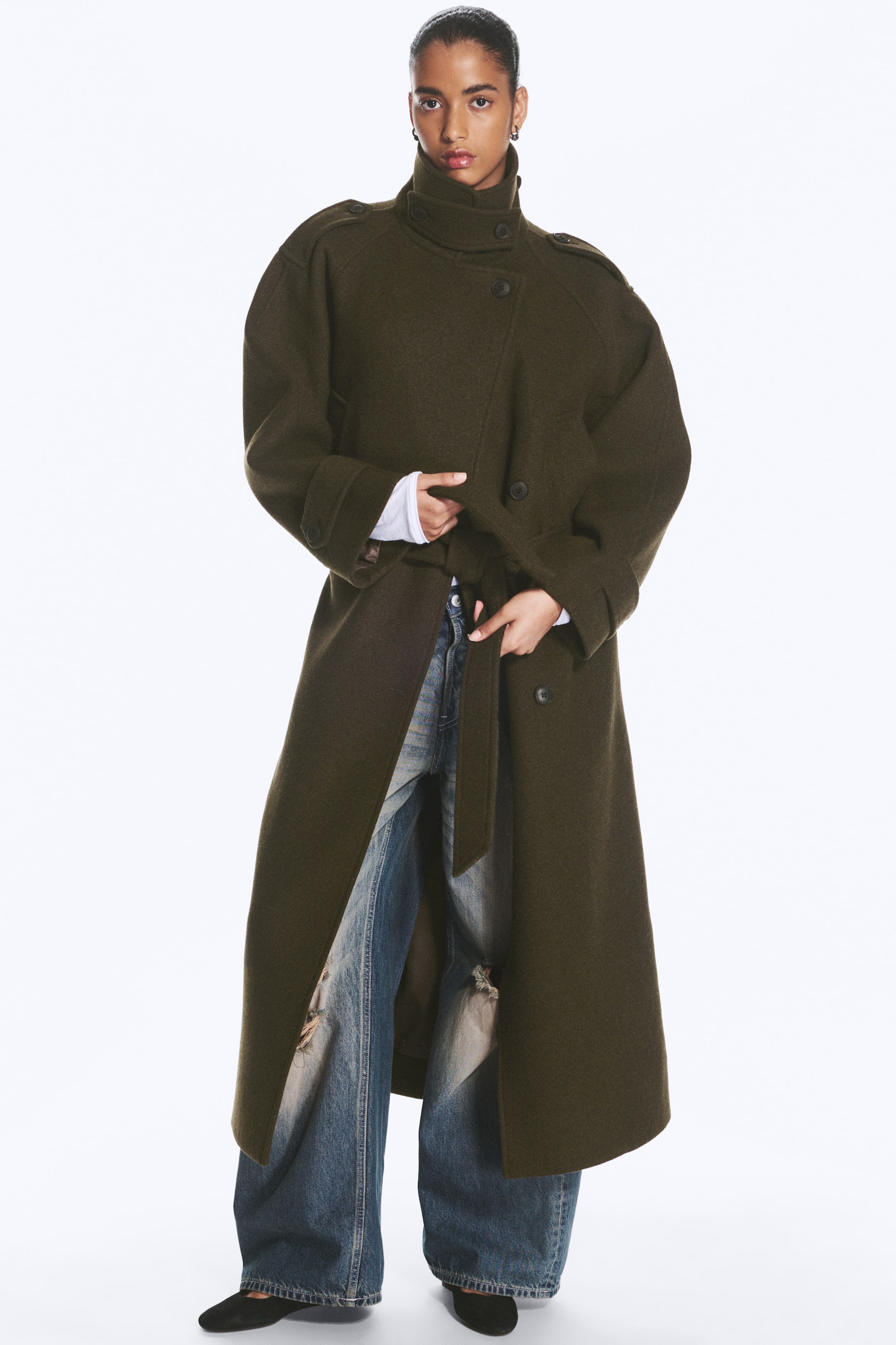 Agrandir l'image: Un long manteau en mélange de laine vert olive, ceinturé, est porté par le mannequin, qui ajuste la ceinture assortie à nouer à la taille. Le manteau présente un col cheminée montant, des pattes d’épaule et une fermeture à boutons, et il est porté avec un jean large effet usé.