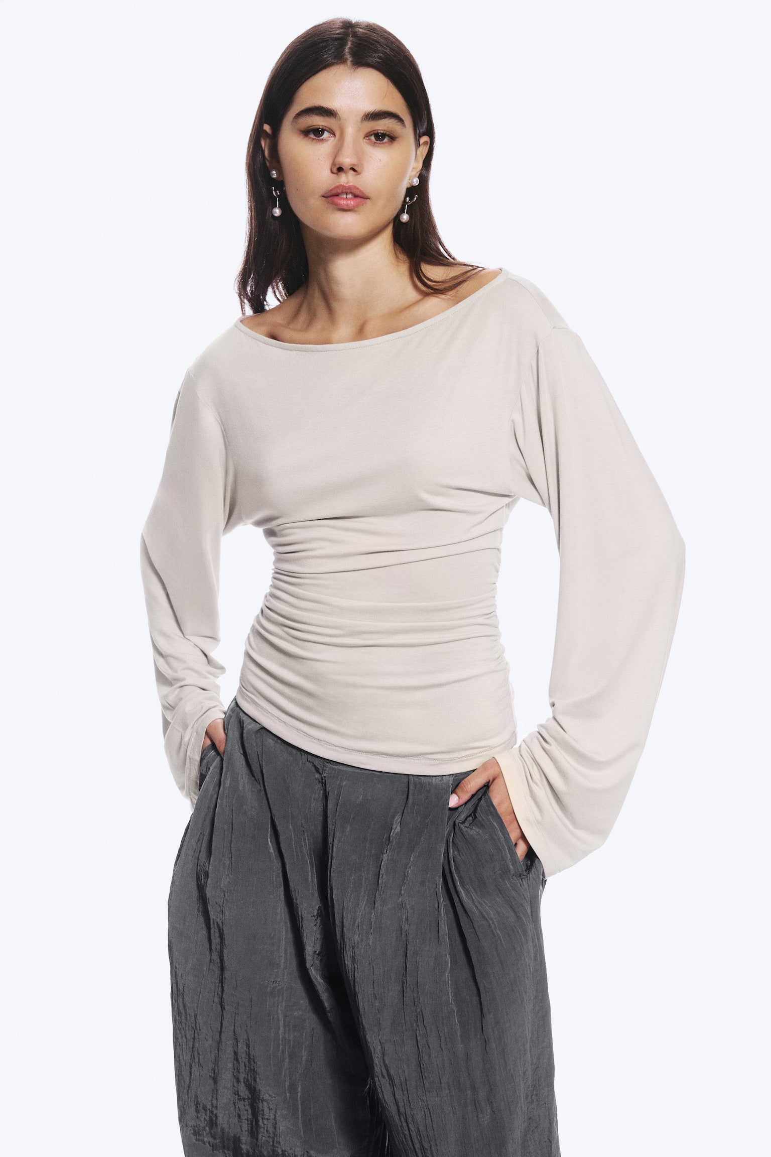 La modelo lleva un top beige claro de manga larga con escote barco, mangas anchas y cintura entallada fruncida. La modelo combina el top con unos pantalones de textura en color gris oscuro, mientras descansa las manos en los bolsillos. - 1