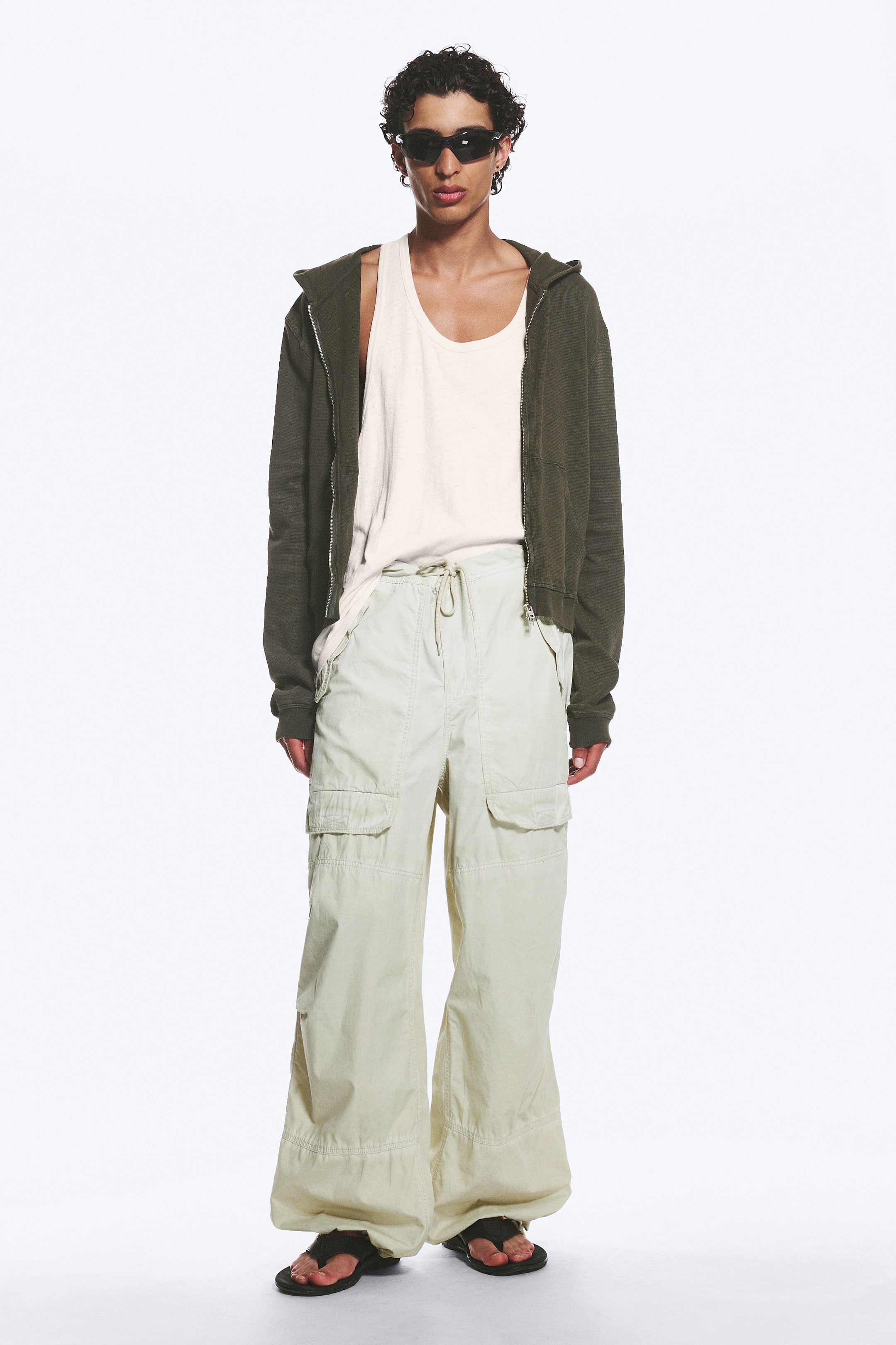 Agrandir l'image: Une tenue superposée est présentée, mettant en vedette un pantalon cargo beige clair à jambes larges avec des poches à rabat et une taille à cordon, qui s'étale sur des sandales noires de style tong. Un hoodie vert olive à fermeture éclair complète est porté ouvert sur un débardeur crème à encolure dégagée, rentré dans le pantalon. Des lunettes de soleil noires enveloppantes sont visibles.