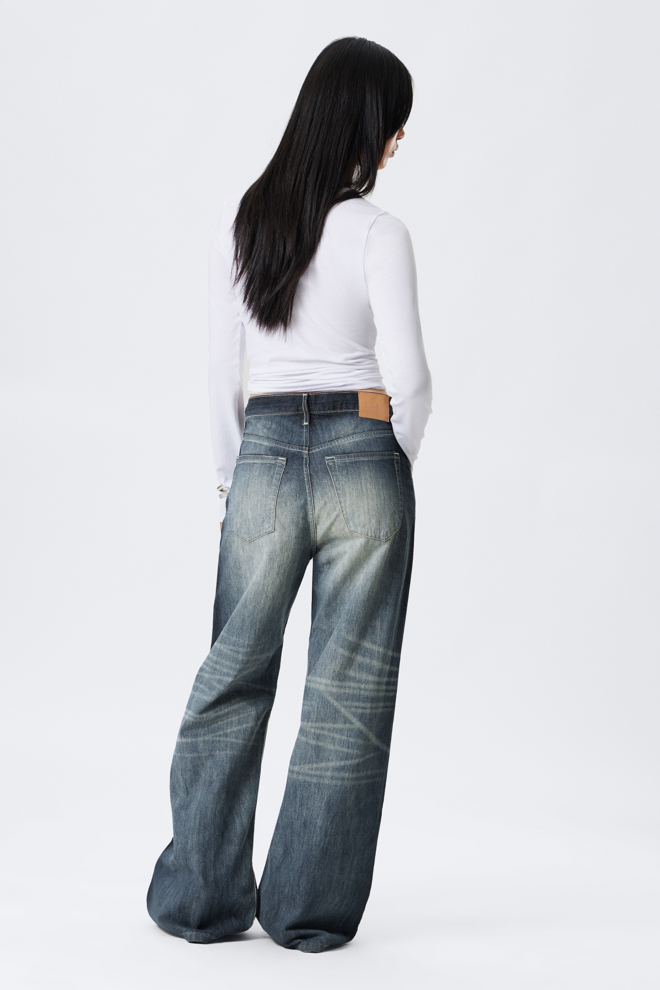 View larger image: Jeans are being shown from the back, featuring a dark wash with faded and distressed areas, including lighter whiskering patterns. They are presenting a wide-leg fit that is pooling at the bottom, and a tan leather patch is visible on the back right waistband.