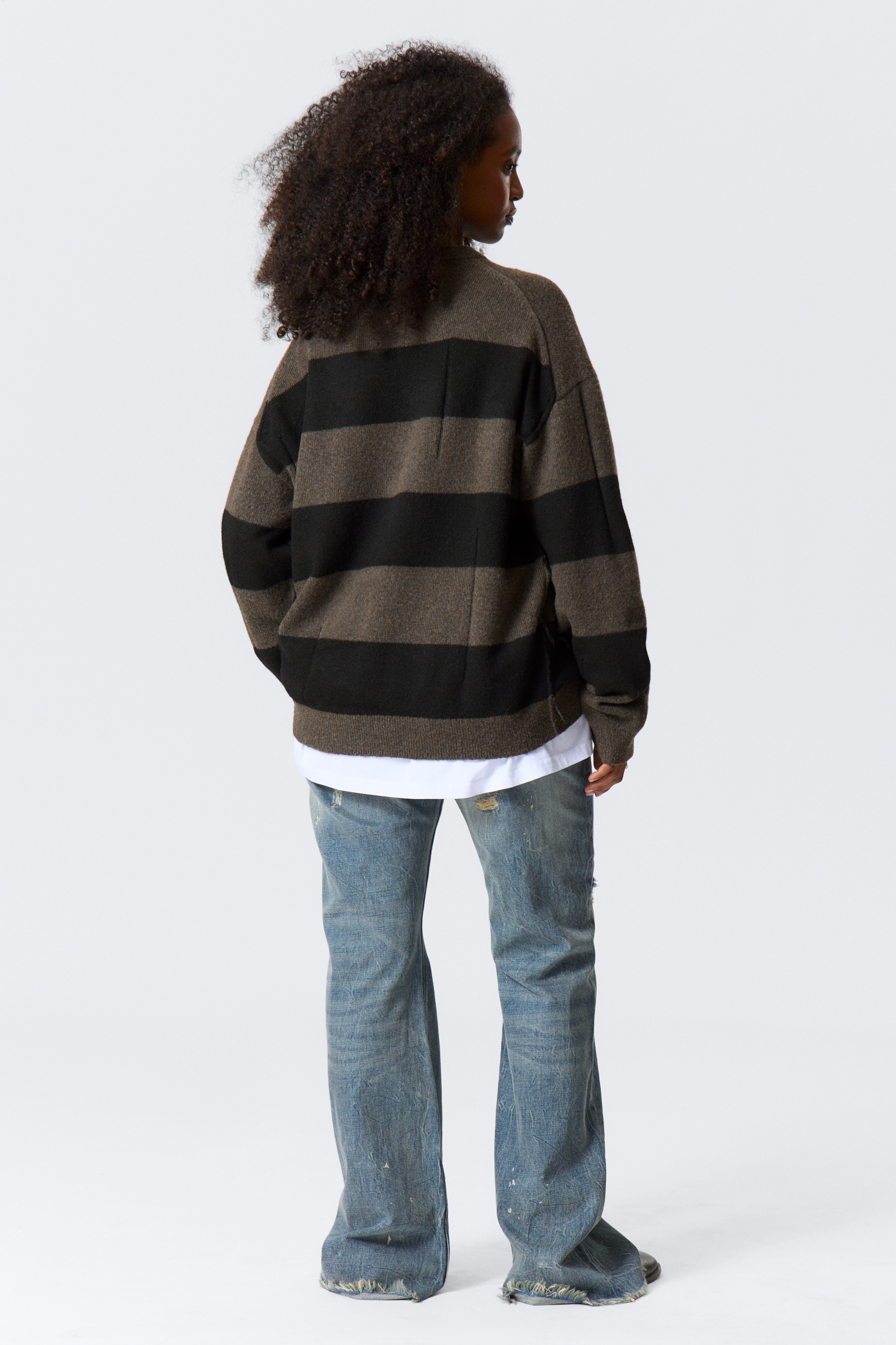 Visualizza immagine più grande: Un maglione a righe oversize, visto da dietro, mostra ampie righe orizzontali in oliva scuro e nero con un orlo a coste. È stratificato su una t-shirt bianca, visibile all'orlo, ed è abbinato a jeans larghi e sbiaditi di colore azzurro.