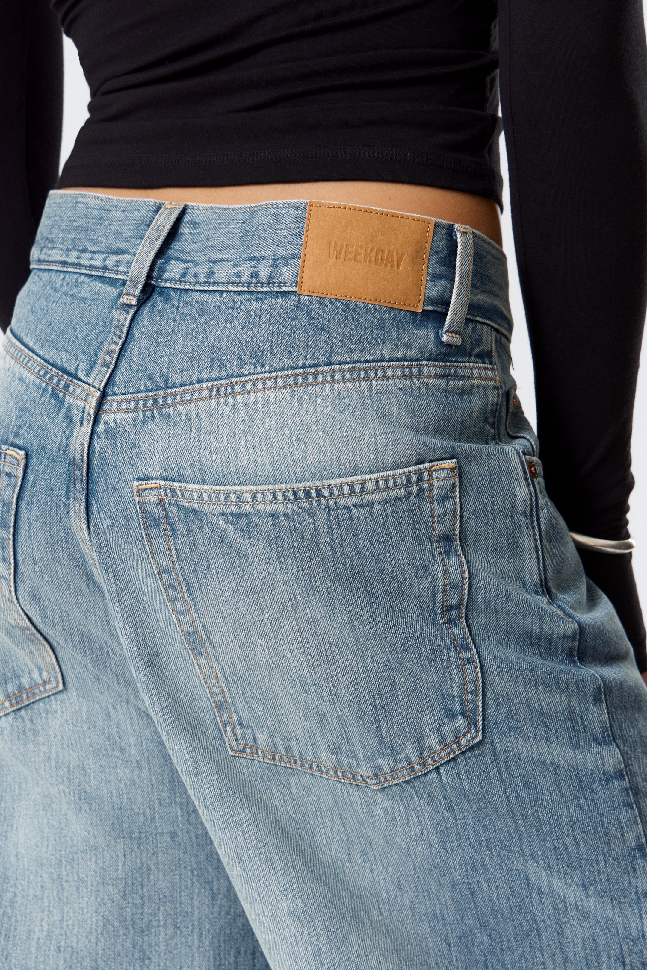 View larger image: Light blue, washed denim jeans are being viewed from the back, angled slightly to the side, revealing a low-rise fit. A brown leather patch embossed with "WEEKDAY" is centered on the back waistband. Two faded patch pockets are visible on the back, detailed with contrast stitching.