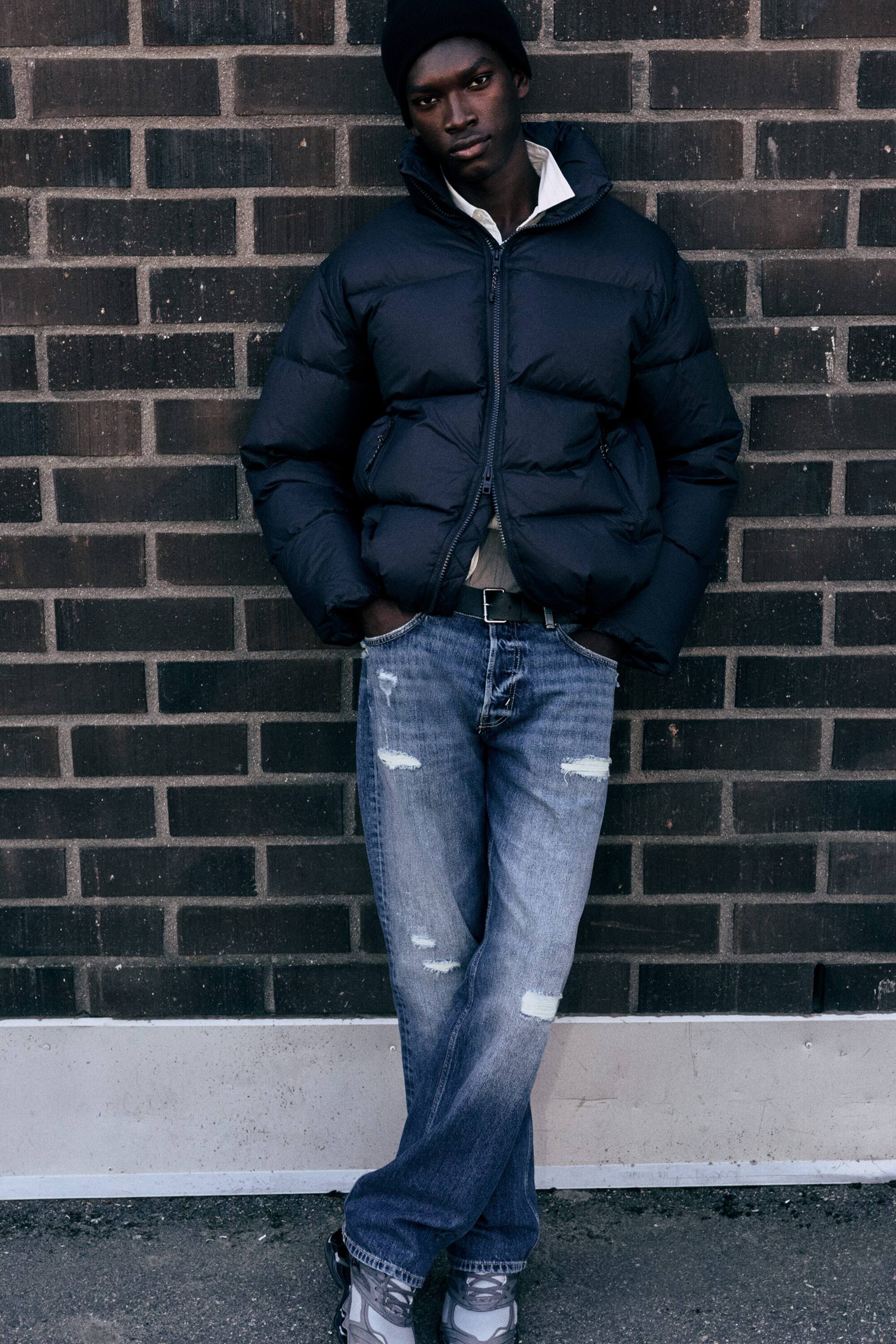 View larger image: A dark navy puffer jacket featuring horizontal quilting, a high stand collar, and a front zip fastening is being worn by the model. The jacket is layered over a white collared shirt and styled with distressed blue jeans.