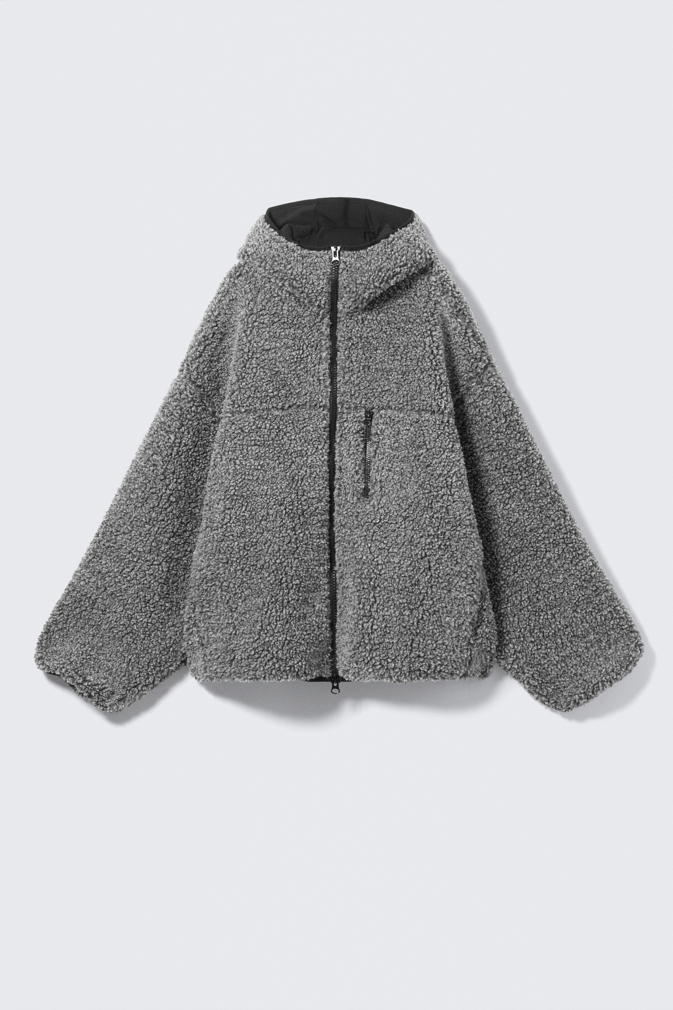 Agrandir l'image: Le devant d'une veste en polaire grise à capuche est exposé. Elle présente un matériau en polaire texturé et bouclé, une doublure noire visible à l'intérieur de la capuche, une fermeture éclair noire à l'avant et une seule poche poitrine zippée noire.