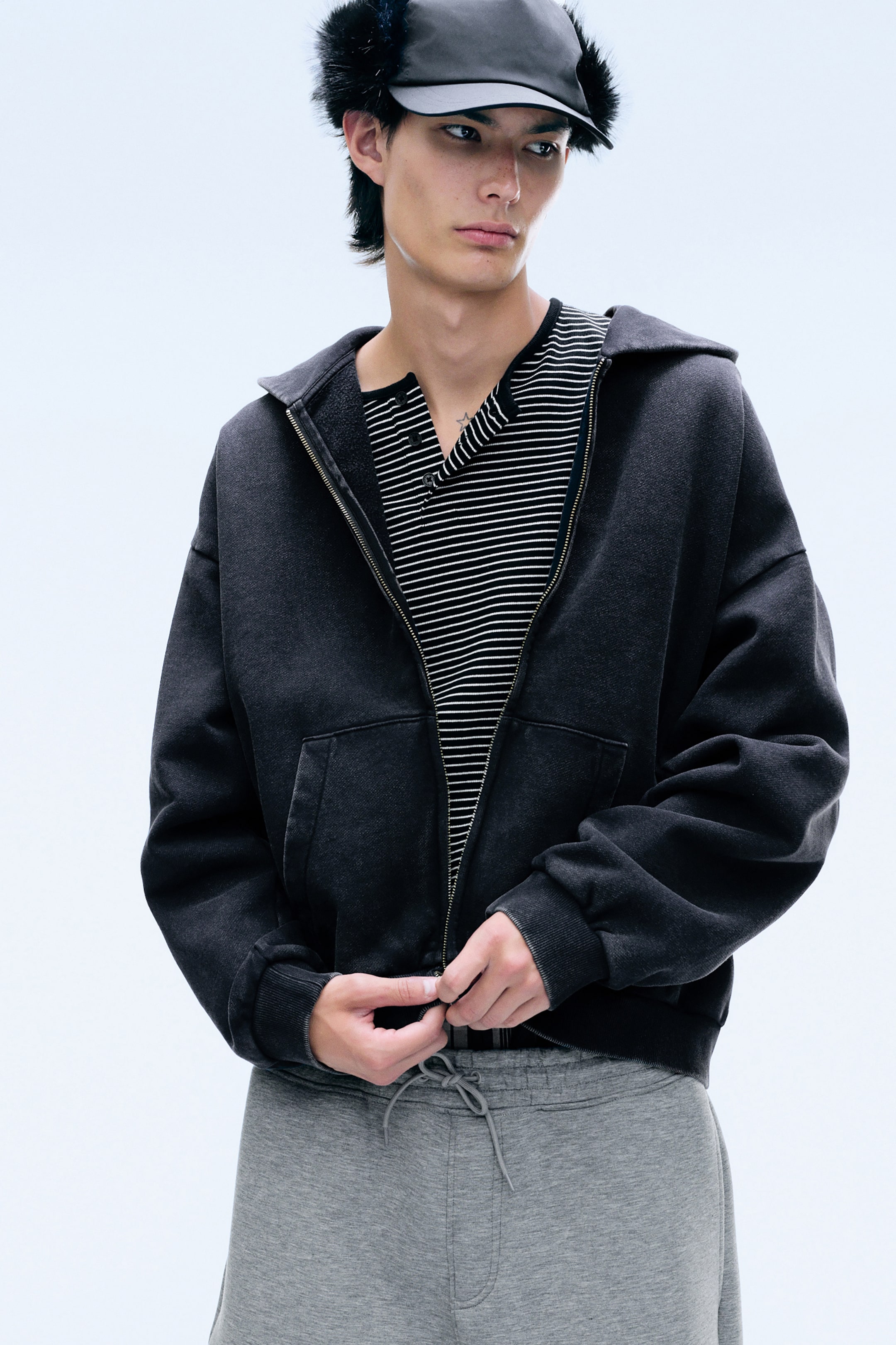 Agrandir l'image: Un hoodie zippé noir délavé est porté par le mannequin, qui ajuste la fermeture éclair à l’ourlet des deux mains. Ce hoodie à coupe ample présente des épaules tombantes, des poches avant et des poignets côtelés, et est superposé à une chemise henley rayée.