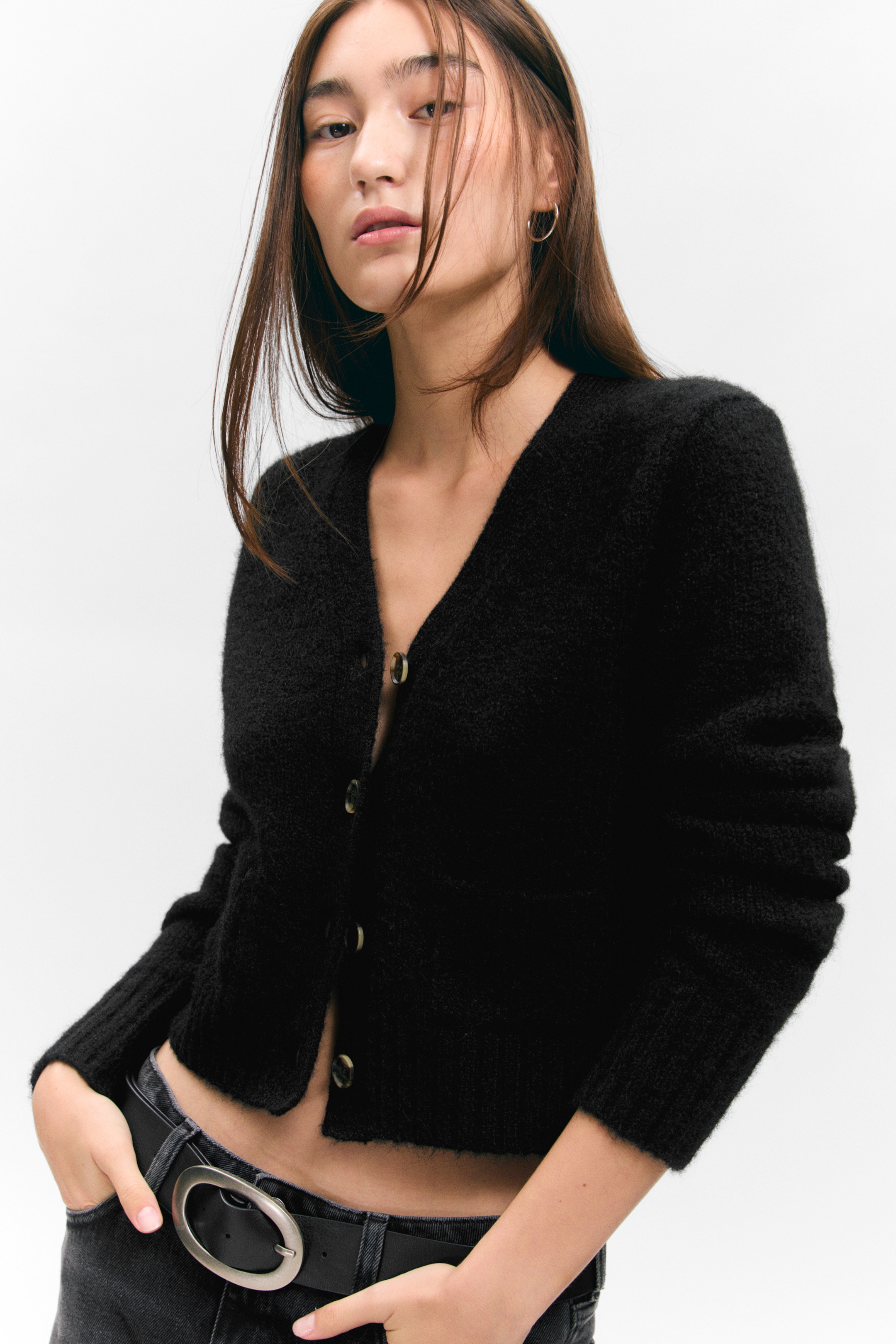 Größeres Bild ansehen: Eine schwarze flauschige Strickjacke ist mit einem V-Ausschnitt und einem Knopfverschluss mit hellen Knöpfen versehen. Die kurze Strickjacke endet über der Taille, enthüllt den Bauch und wird aus einem frontalen Winkel präsentiert.