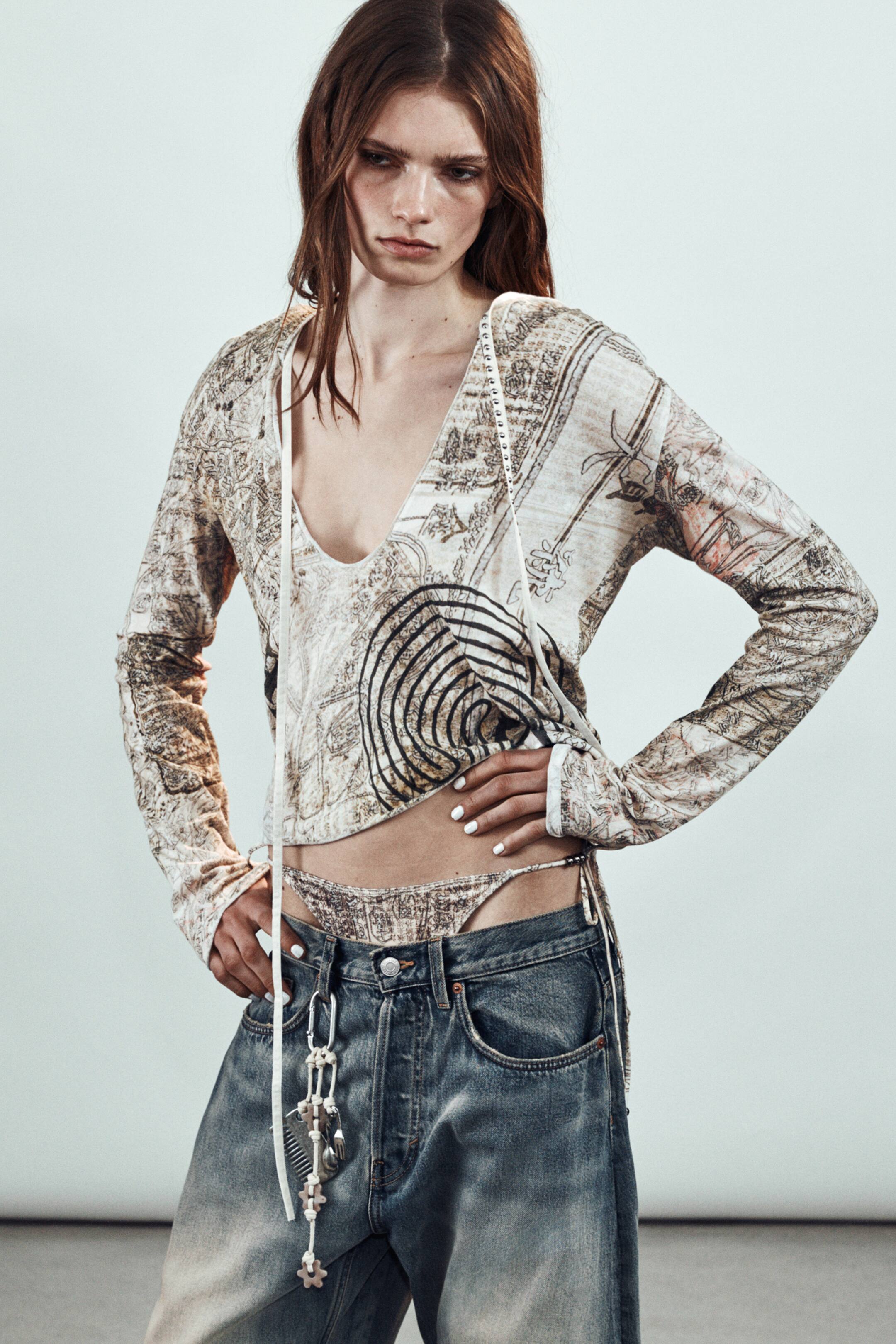 Προβολή μεγαλύτερης εικόνας: Ένα κοντό, μακρυμάνικο τοπ με βαθύ V-λαιμόκοψη και εκτύπωση εμπνευσμένη από παλαιό χάρτη σε μπεζ και καφέ, διαθέτει λευκά κορδόνια κατά μήκος της λαιμόκοψης. Ένα ασορτί στρινγκ με το ίδιο μοτίβο είναι εμφανώς στυλιζαρισμένο πάνω από τη ζώνη χαμηλοκάβαλων τζιν.