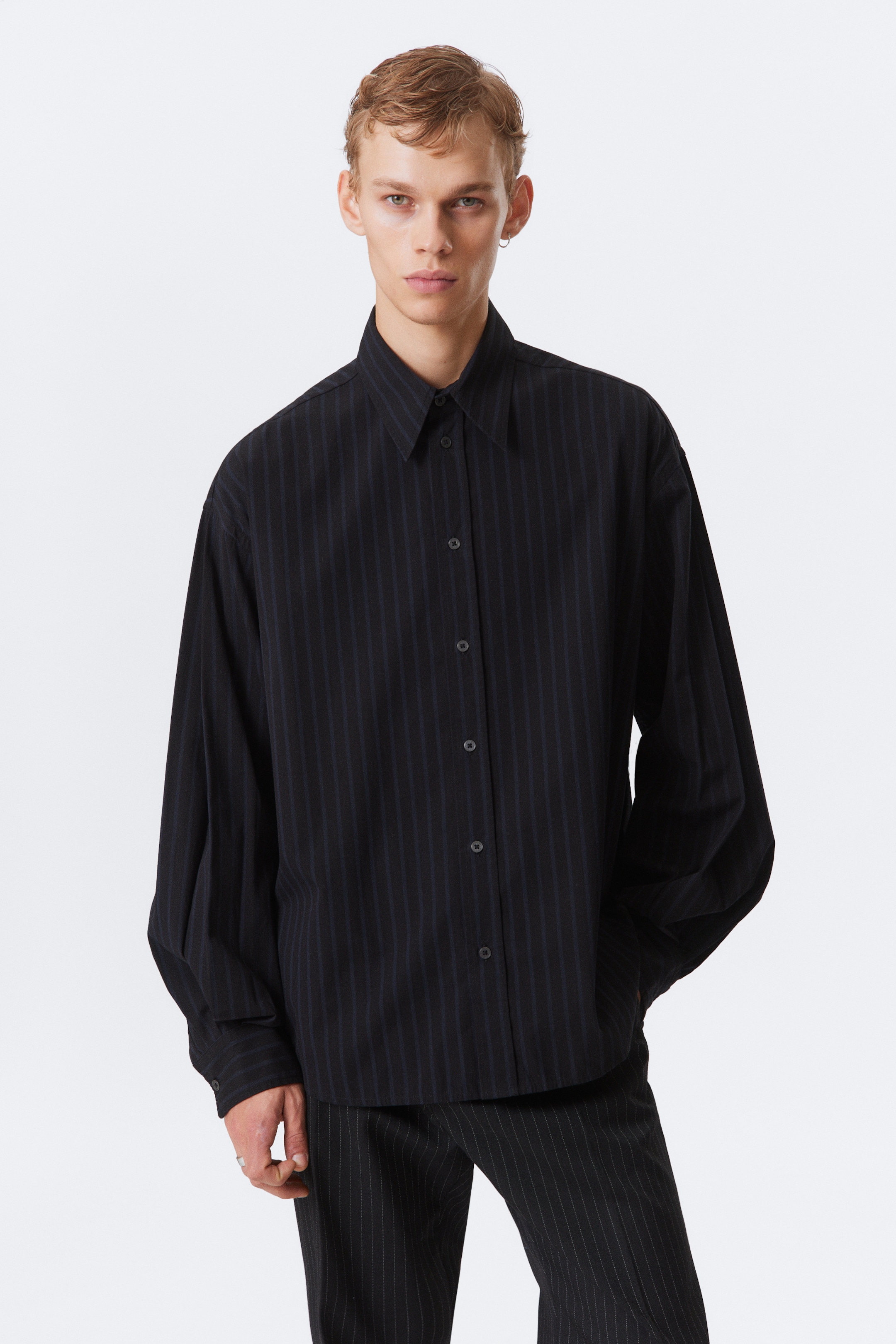 Ver imagen más grande: Una camisa negra presenta sutiles rayas oscuras verticales a lo largo de su tela. Está diseñada con un clásico cuello en punta, una tapeta de botones frontal con botones oscuros y mangas largas y sueltas con puños abotonados.