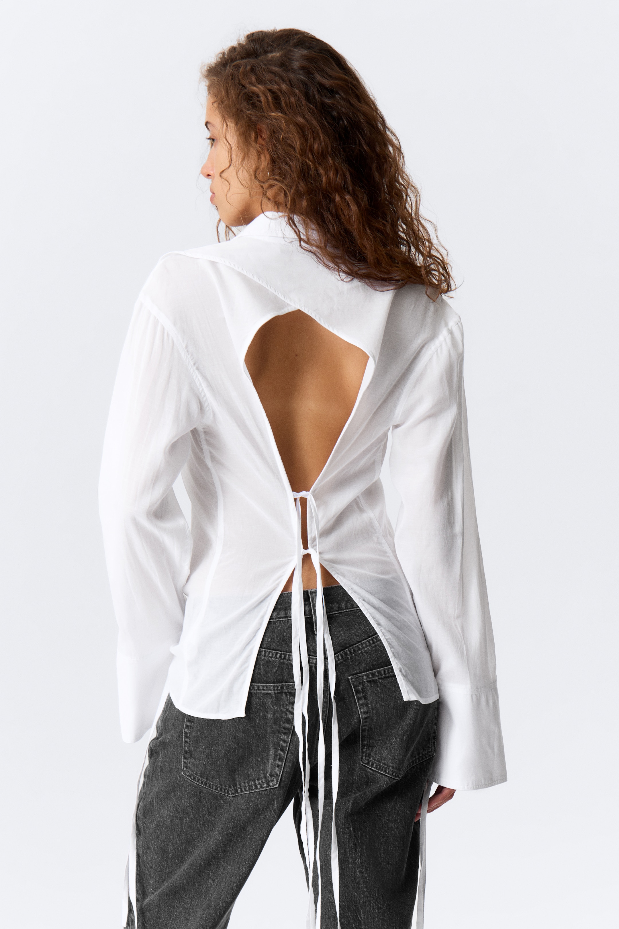 Ver imagem maior: Uma camisa branca está a ser apresentada de costas, com um grande recorte que revela a parte central das costas. As fitas de tecido branco estão a apertar a cintura, criando uma bainha dividida que se estende sobre umas jeans de cor escura, e a camisa é finalizada com mangas compridas e punhos largos.