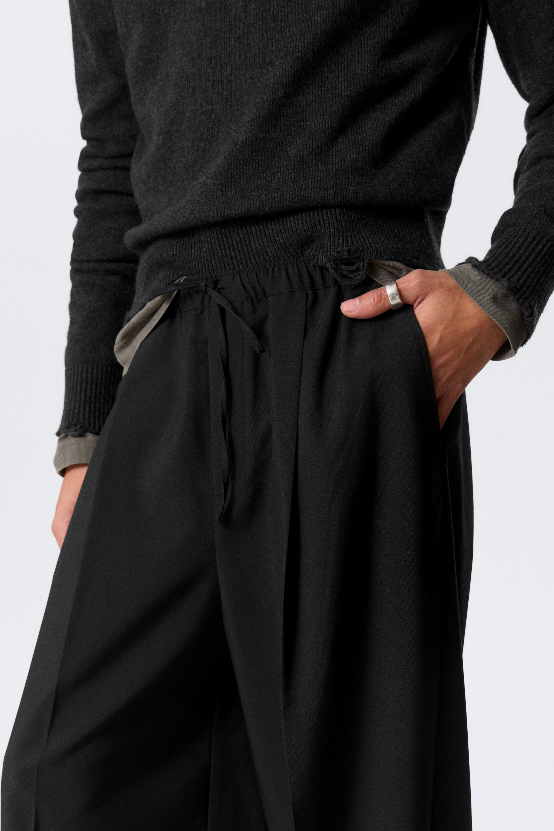Grotere afbeelding bekijken: Zwarte volgesneden broek wordt getoond vanuit een frontaal perspectief, met een elastische tailleband met trekkoord, zachte plooien en zijzakken. Een donkergrijze trui wordt over de tailleband gelaagd, met een hand die in een van de broekzakken rust.