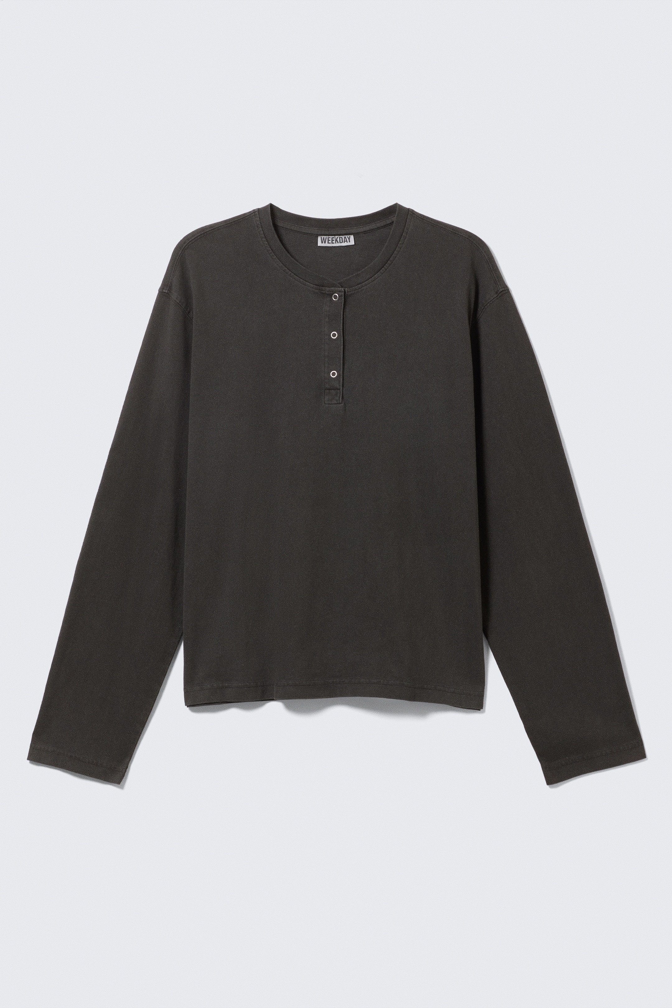 Visa större bild: En mörkgrå långärmad Henley-tröja presenteras framifrån, med en rund halsringning och en treknappslå. Den visar långa ärmar och en rak fåll, medan en "WEEKDAY"-etikett är synlig på baksidan av nacken.
