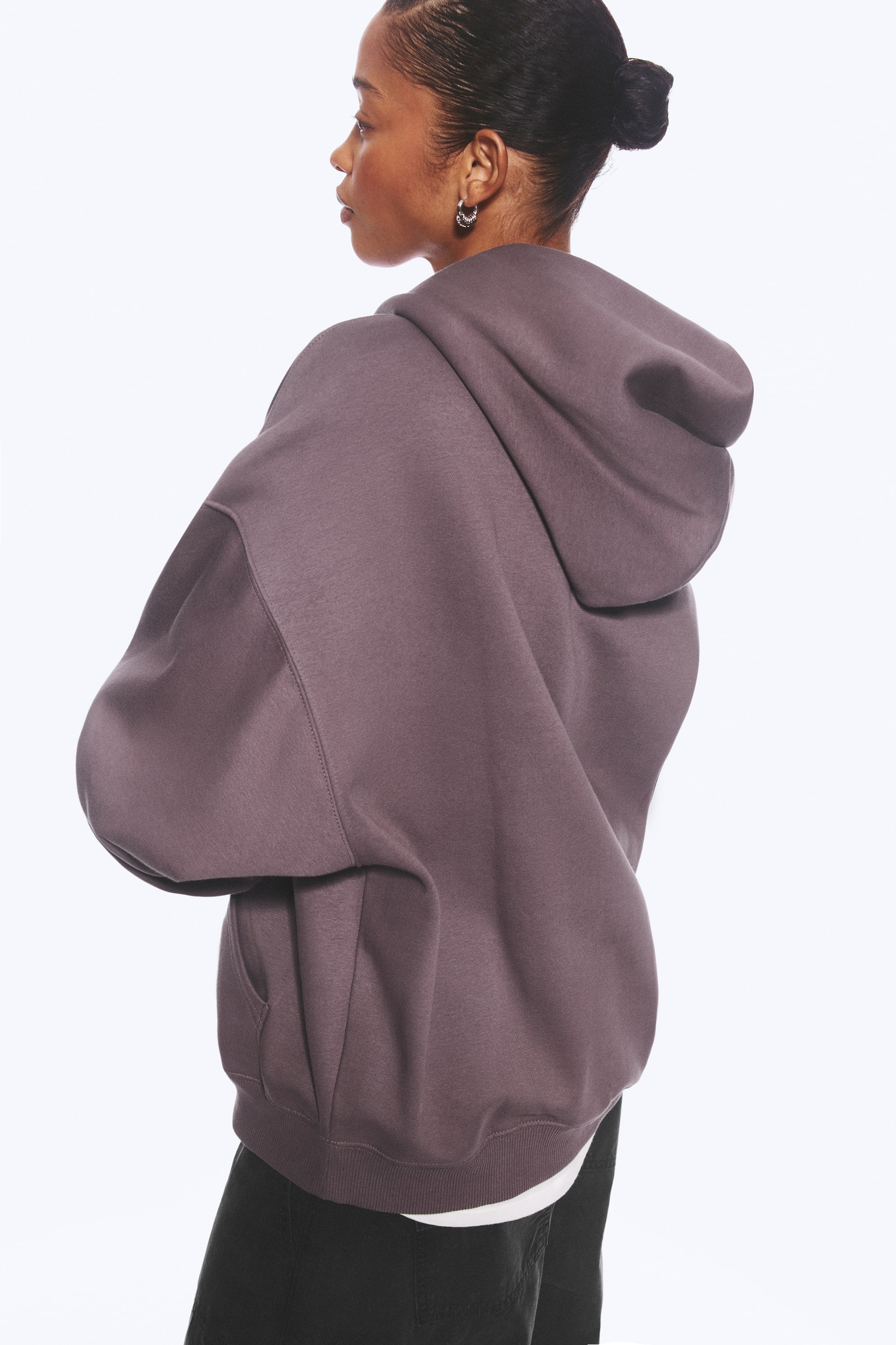Visa större bild: En oversized hoodie i dämpad lila bärs av modellen och visas i trekvarts bakifrån. Hoodien har en avslappnad silhuett med sänkta axlar, en stor huva och en ribbstickad fåll, med en vit undertröja som syns undertill.