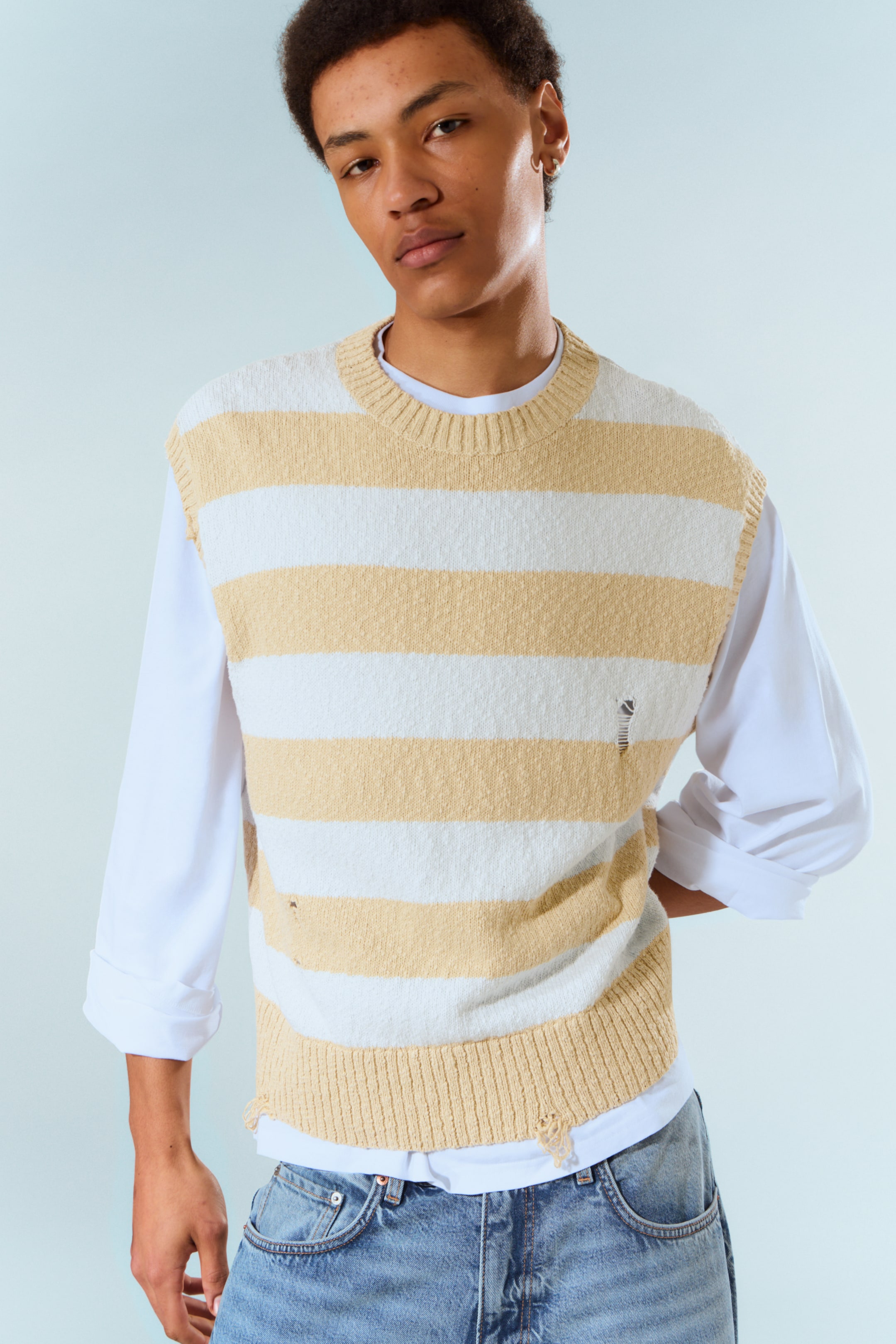 Visa större bild: En tröjväst med horisontella ränder i växlande beige och vitt, med ribbad rund hals, ribbade ärmhål och en ribbad fåll med slitna detaljer på kroppen och längs nederkanten. Den bärs över en vit långärmad skjorta.