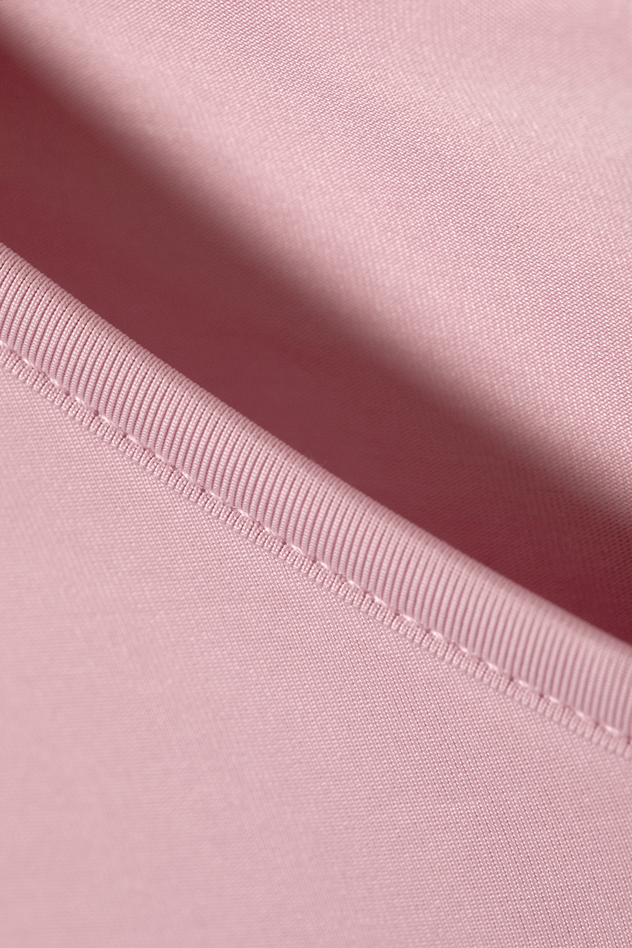Größeres Bild ansehen: Ein rosa Stoff zeigt eine glatte Strickstruktur neben einem gerippten Strickbesatz. Eine Linie aus weißer Naht verbindet sichtbar die beiden unterschiedlichen Abschnitte, wobei eine sanfte Falte Tiefe im Stoff erzeugt.