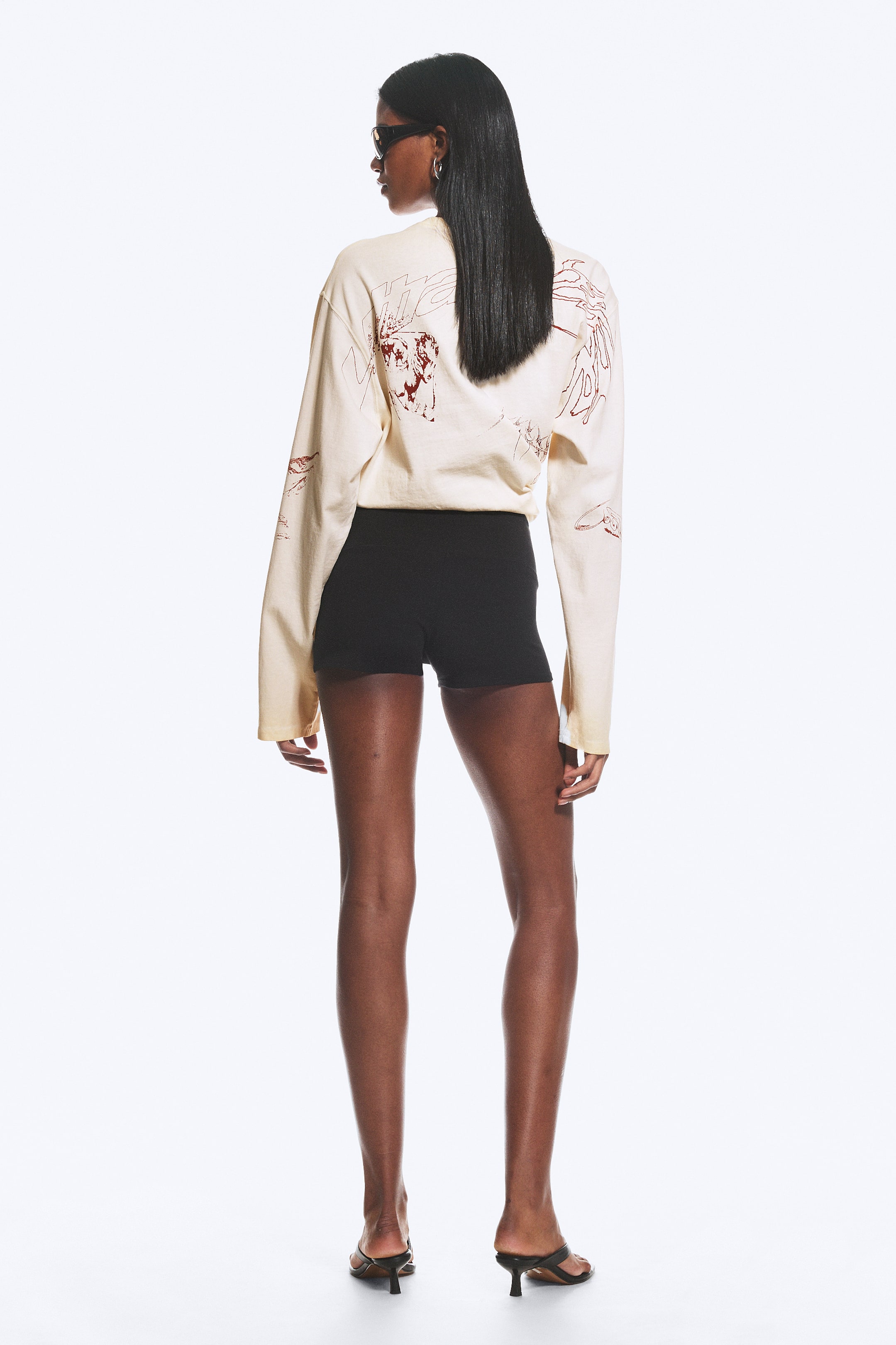 Größeres Bild ansehen: Ein cremefarbenes Langarmshirt mit roten abstrakten Grafiken auf dem Rücken und den Ärmeln wird über einer schwarzen, eng anliegenden High-Waist-Shorts getragen. Das Oberteil zeigt einen lockeren, oversized Schnitt, während die Shorts eng am Körper anliegen. Die Kleidungsstückkombination ist vom Betrachter abgewandt.