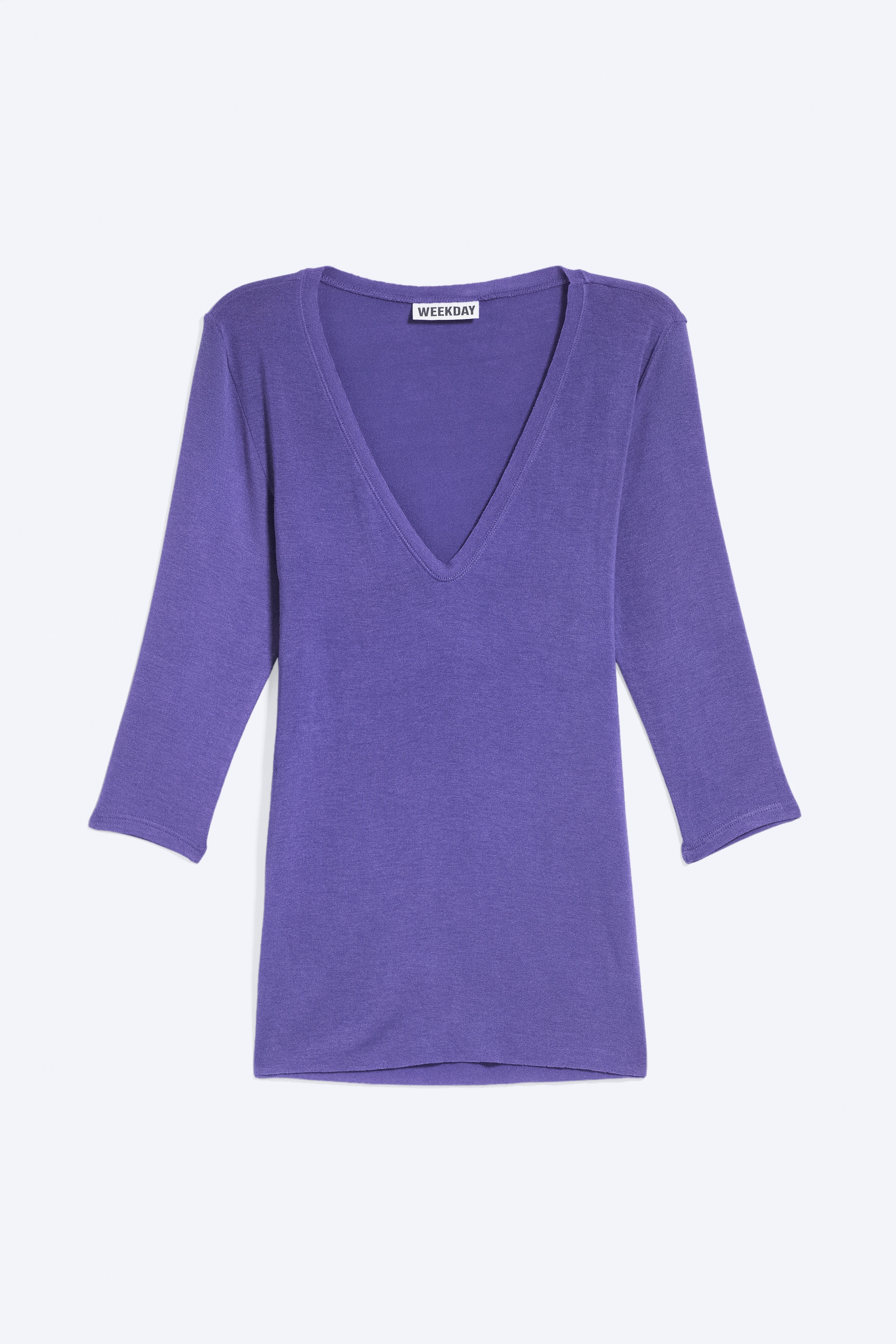 Ein figurbetontes Oberteil in Violett mit Dreiviertelärmeln und tiefem, rundem Ausschnitt wird vom Model getragen. Das Oberteil ist mit ausgewaschenen blauen Jeans kombiniert, wobei das Model die Hände in den vorderen Taschen hat.