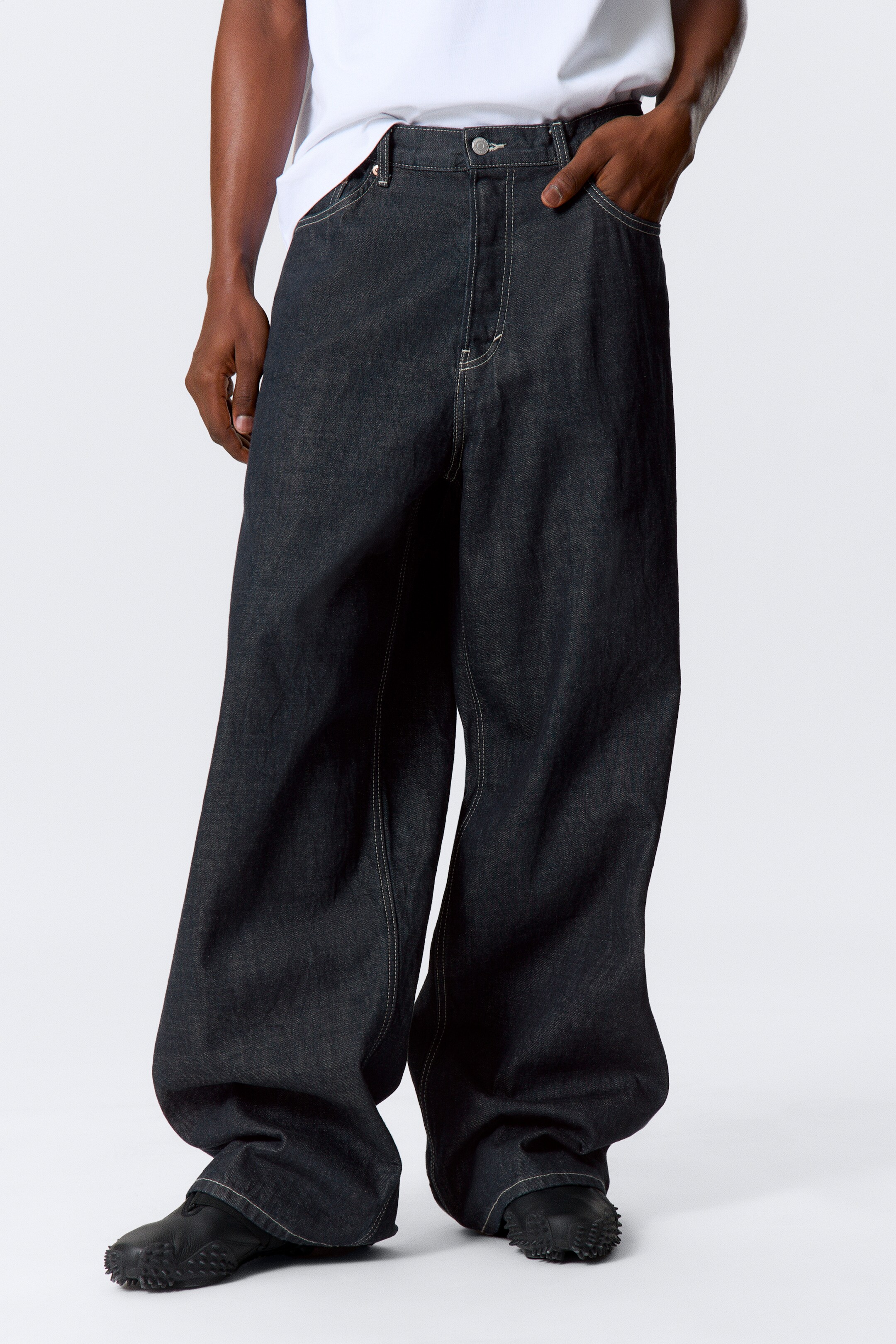 Grösseres Bild ansehen: Dunkel gewaschene, weit geschnittene Jeans zeichnen sich durch weiße Kontrastnähte entlang der Nähte, Taschen und des Bunds aus. Die Jeans weisen einen sehr lockeren, lässigen Sitz auf, wobei die weiten Beine über den schwarzen Schuhen des Trägers zusammenfallen, da sie aus einem leicht angedeuteten Frontalwinkel präsentiert werden.