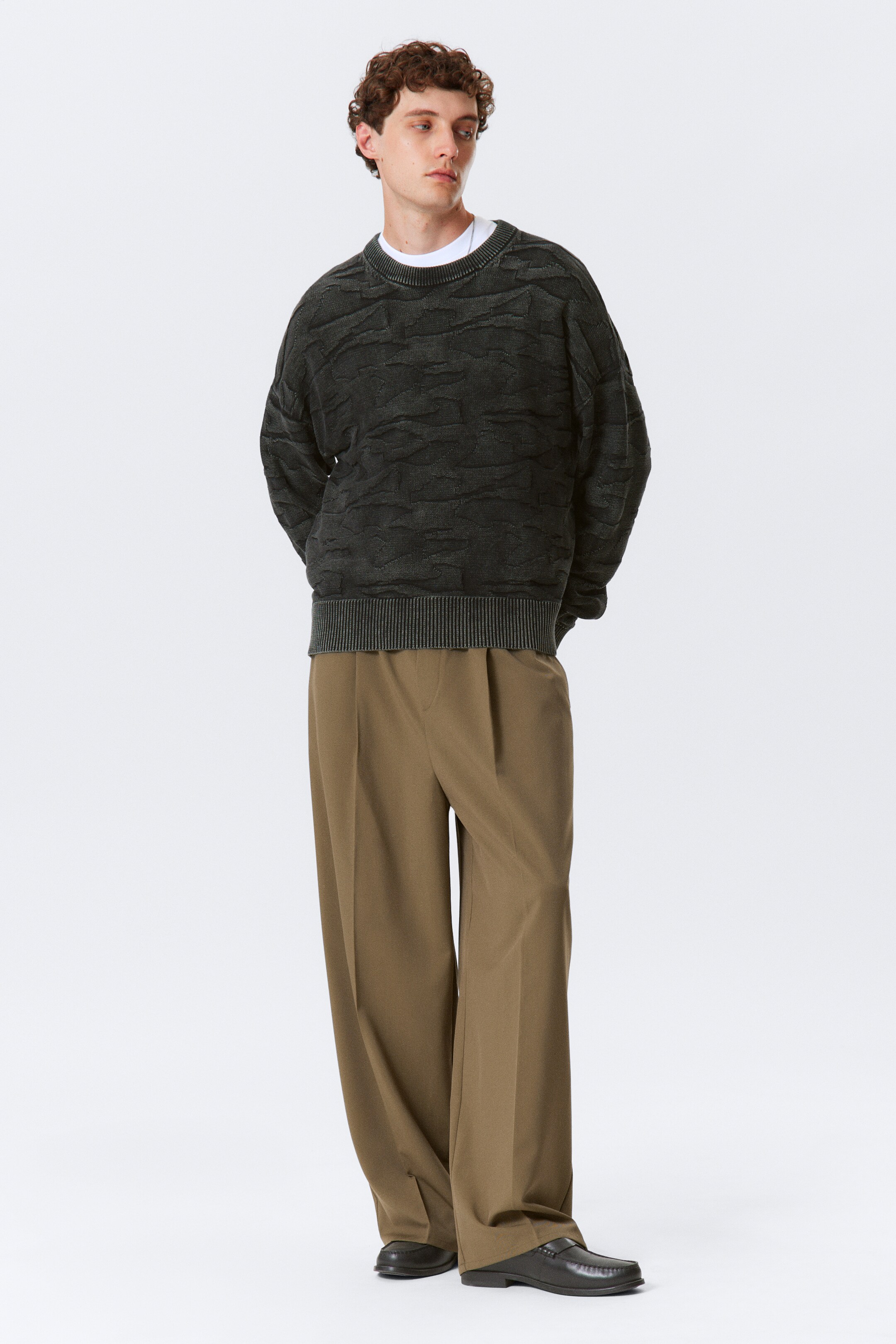 Grotere afbeelding bekijken: Een donkergrijze crewneck trui, met een reliëf van een gekreukte textuur en een geribbelde kraag en zoom, wordt gedragen in een losse, oversized pasvorm over een wit t-shirt. Het wordt gestyled met wijde, geplooide olijfbruine broeken en donkere loafers, met de volledige productpresentatie zichtbaar vanuit een vooraanzicht.