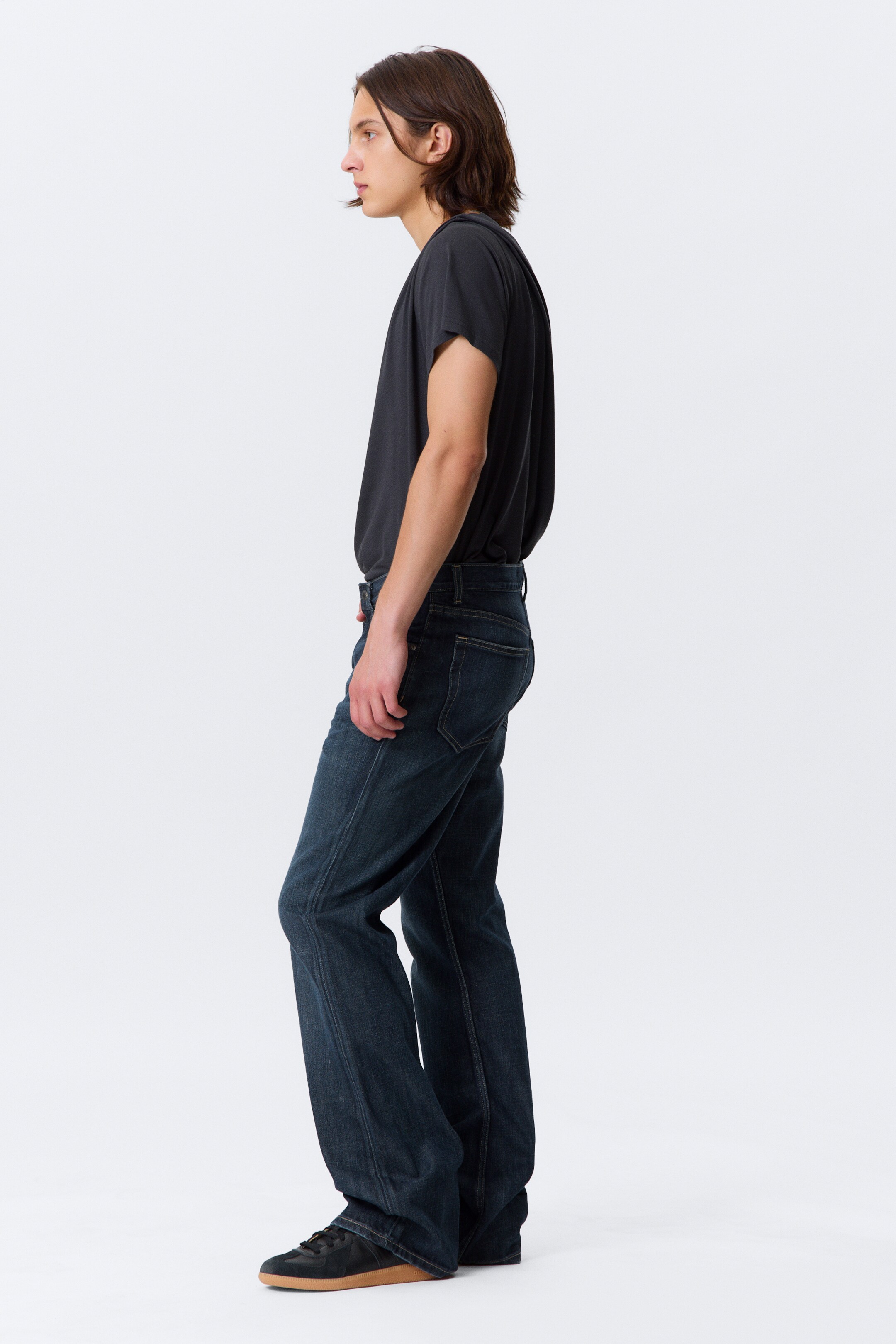 View larger image: Dark wash denim jeans are being worn by a person facing left, showcasing a relaxed straight-leg fit with visible stitching and a back pocket, resting over dark sneakers. A loose-fitting dark t-shirt with short sleeves is being worn, tucked into the jeans.