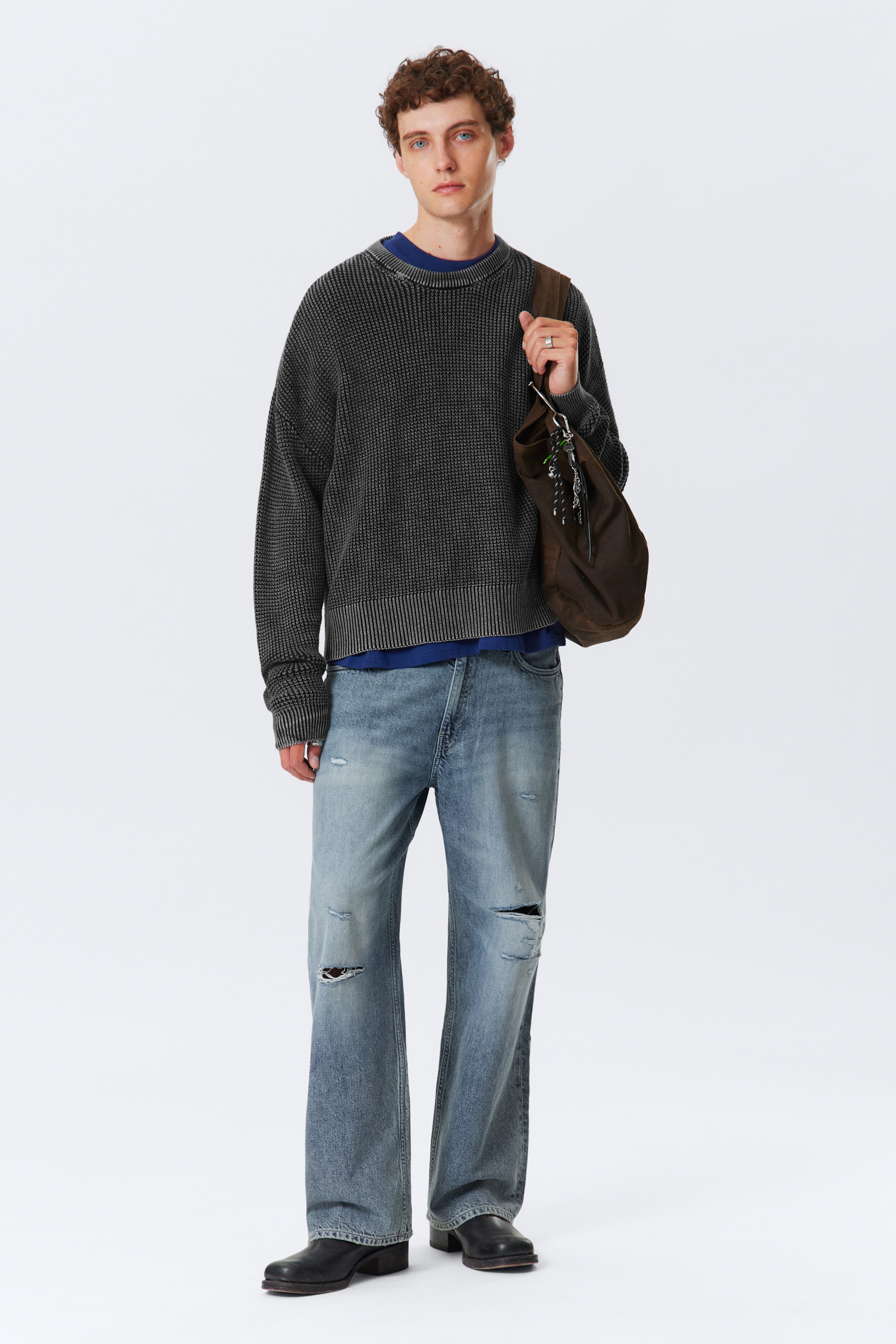 Größeres Bild anzeigen: Ein dunkelgrauer Waffelstrickpullover mit geripptem Rundhalsausschnitt, Bündchen und Saum wird in einer Ganzkörperansicht von vorne präsentiert, über einem blauen T-Shirt gestylt. Es wird mit hell gewaschenen, distressed, weit geschnittenen Jeans kombiniert, die Risse an den Knien und Oberschenkeln aufweisen, schwarzen Schuhen mit niedrigem Absatz und einer braunen Schultertasche mit hängenden Charms, die auf der rechten Seite ruht.