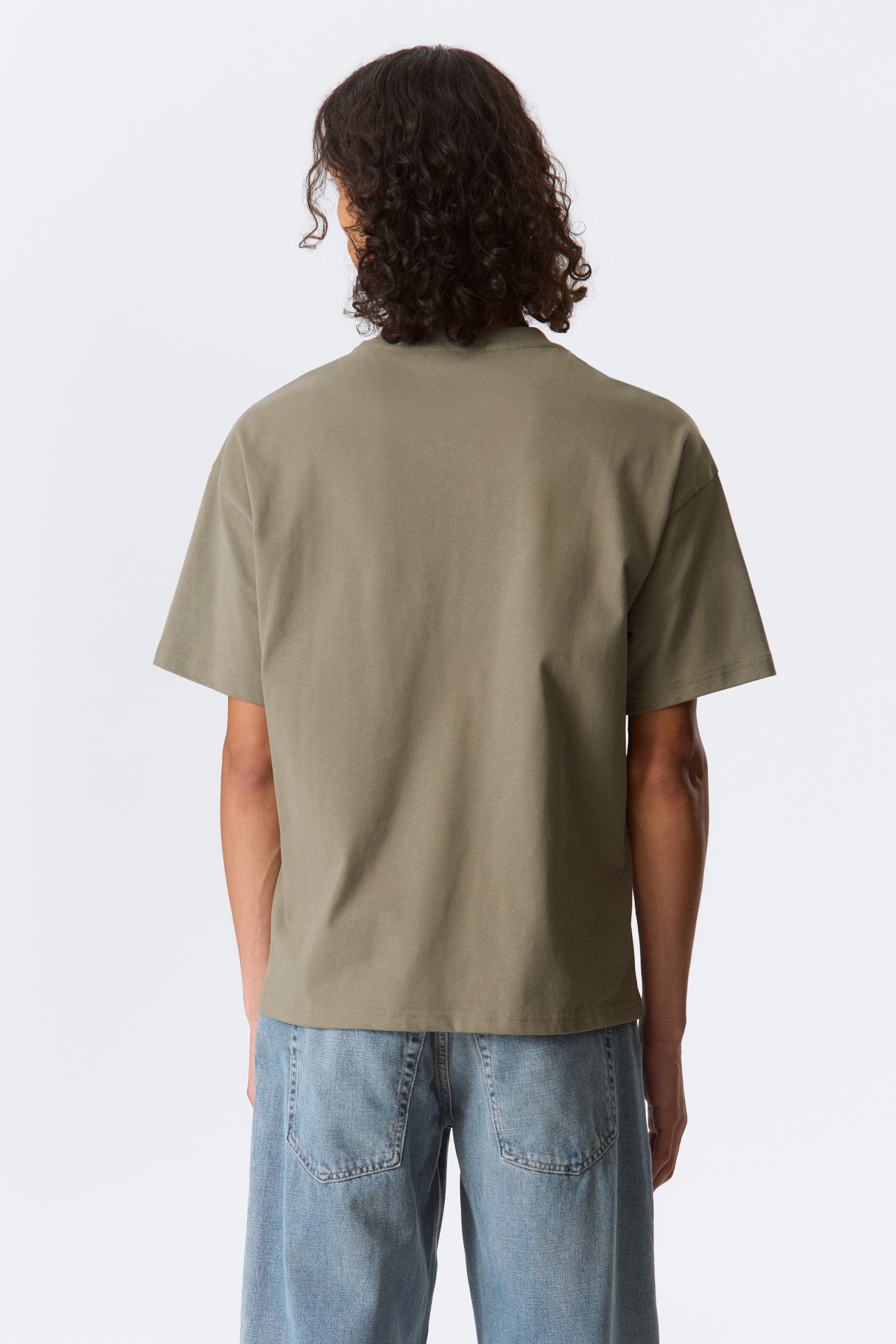 Grotere afbeelding bekijken: Een olijfgroene t-shirt toont de achterkant, met een zichtbare ronde hals, een losse pasvorm en korte, wijde mouwen. De rechte zoom valt over blauwe denim jeans.
