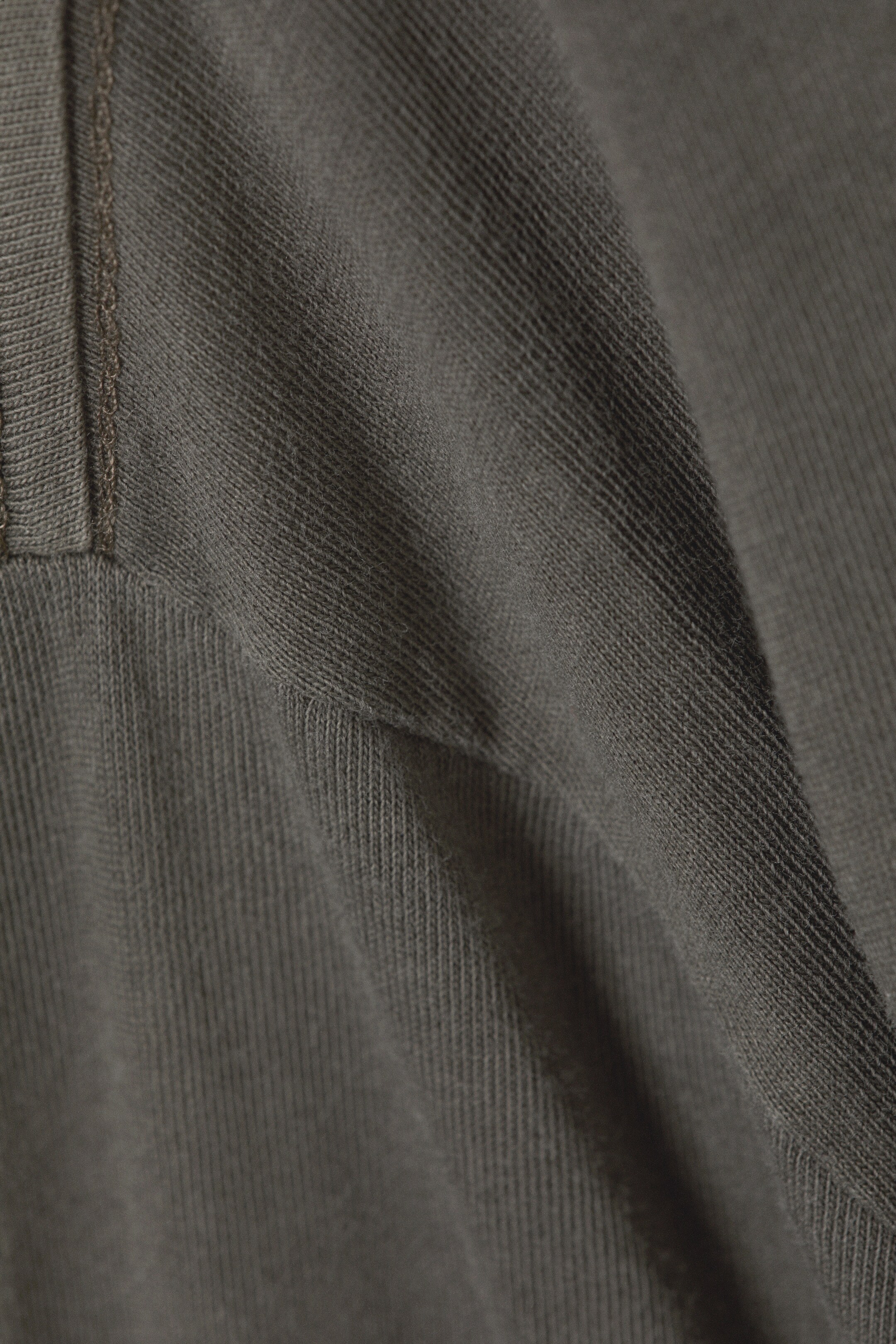 Ver imagen más grande: Una tela de punto acanalada de color verde oliva oscuro presenta una costura visible con dos líneas paralelas de costura tonal. La tela cae suavemente, formando pliegues que acentúan su textura acanalada.