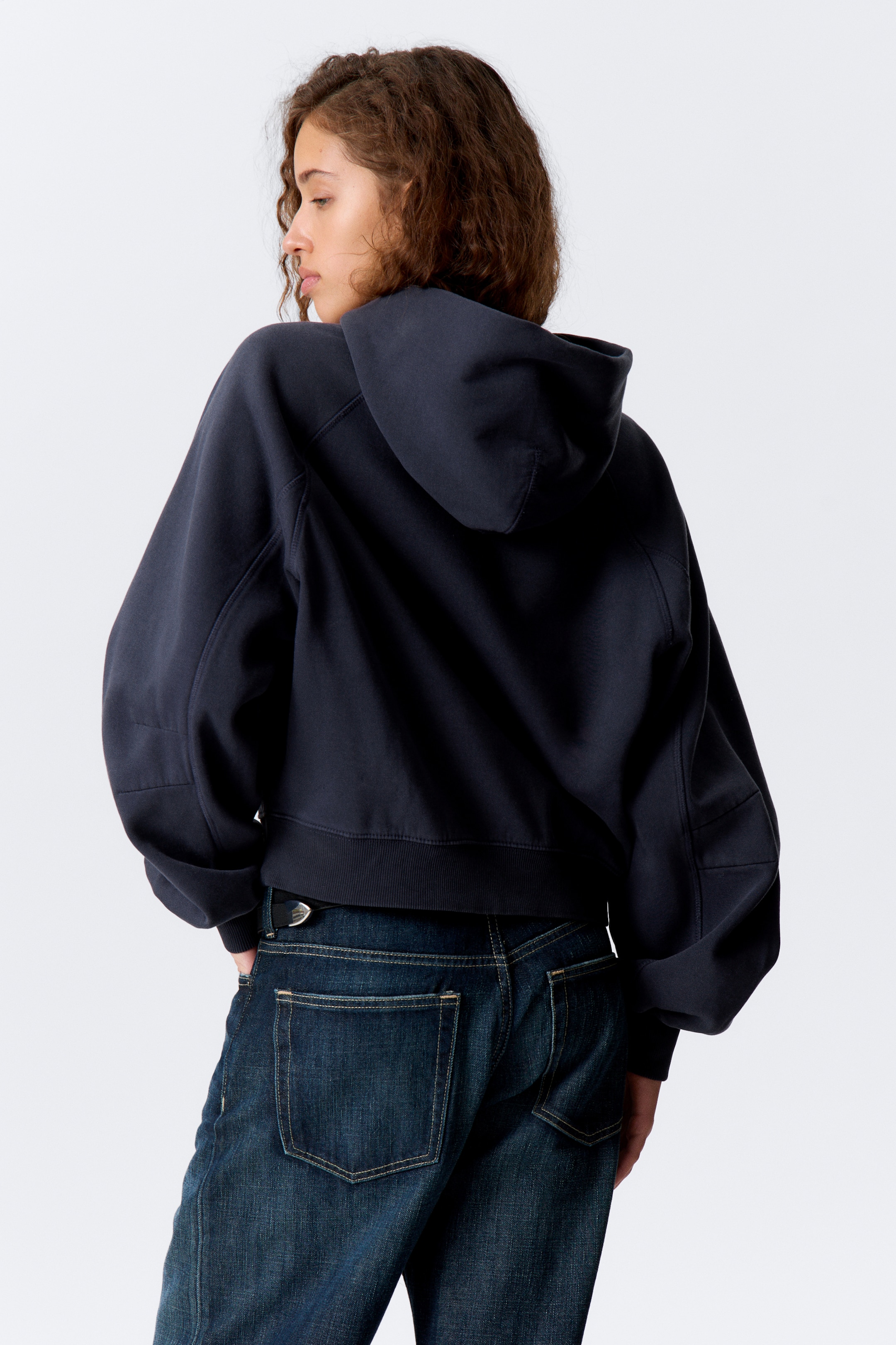 Größeres Bild ansehen: Ein dunkel gefärbter Hoodie wird aus einer rückwärtigen linken Perspektive gezeigt, mit einer Kapuze, voluminösen, gepanelten langen Ärmeln mit gerippten Bündchen und einem gerippten Saum, der an der Taille über dunklen Jeans sitzt.