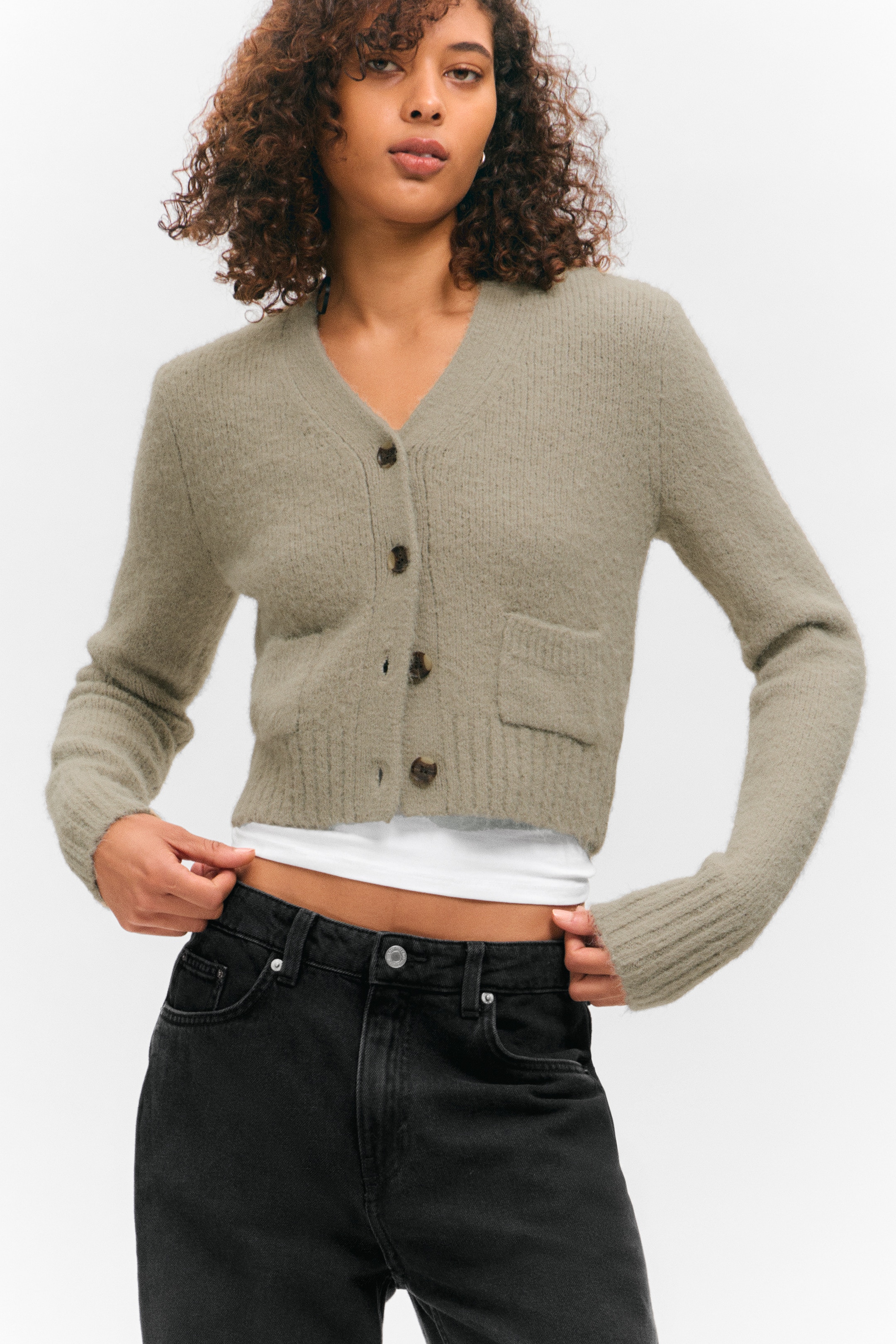 Ingrandisci l'immagine: Un cardigan corto verde oliva chiaro è presentato dal davanti, con una texture a maglia pelosa, uno scollo a V e una chiusura frontale a quattro bottoni con bottoni marmorizzati marroni. Presenta anche una tasca applicata sul lato sinistro di chi lo indossa, insieme a polsini e orlo a coste, mentre è abbinato aperto sopra un top bianco.