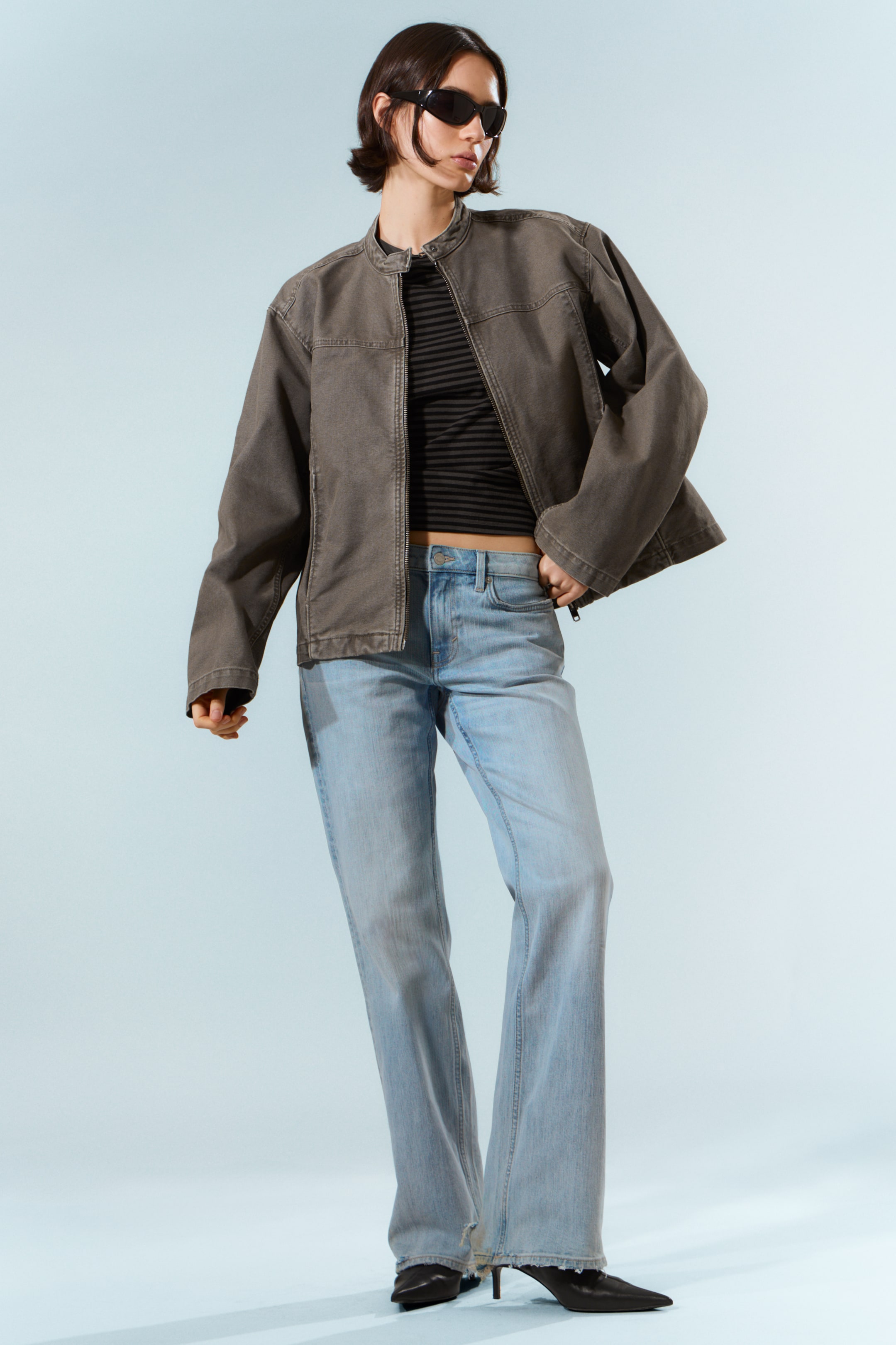 Näytä suurempi kuva: Tummanharmaa pesty käyttöjakku, leikattu lantion pituiseksi, esitetään. Siinä on pystykaulus, täyspitkä vetoketjukiinnitys ja näkyvät saumat olkapäillä ja rinnassa. Kaksi vaakasuuntaista taskua näkyvät vyötäröllä, ja takki on puettu auki mustan ja harmaan raidallisen yläosan päälle.