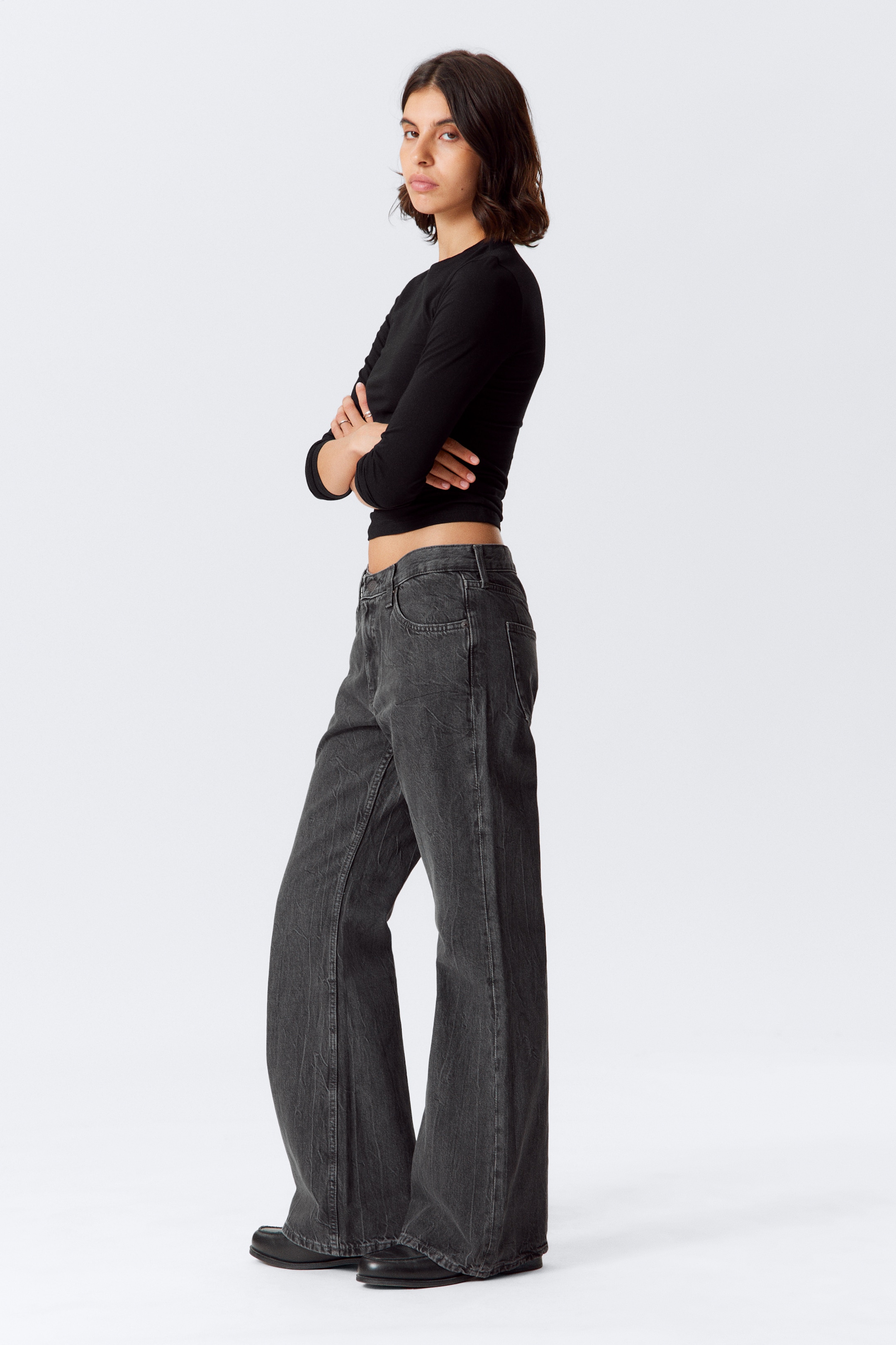 Grösseres Bild ansehen: Ein Paar dunkelgraue Jeans mit weitem Bein, das eine gewaschene Oberfläche und sichtbare Fronttaschen aufweist, wird getragen. Die Jeans sind mit einem schwarzen, langärmeligen, figurbetonten, bauchfreien Oberteil gestylt, das einen Rundhalsausschnitt hat und den Bauch freilegt.