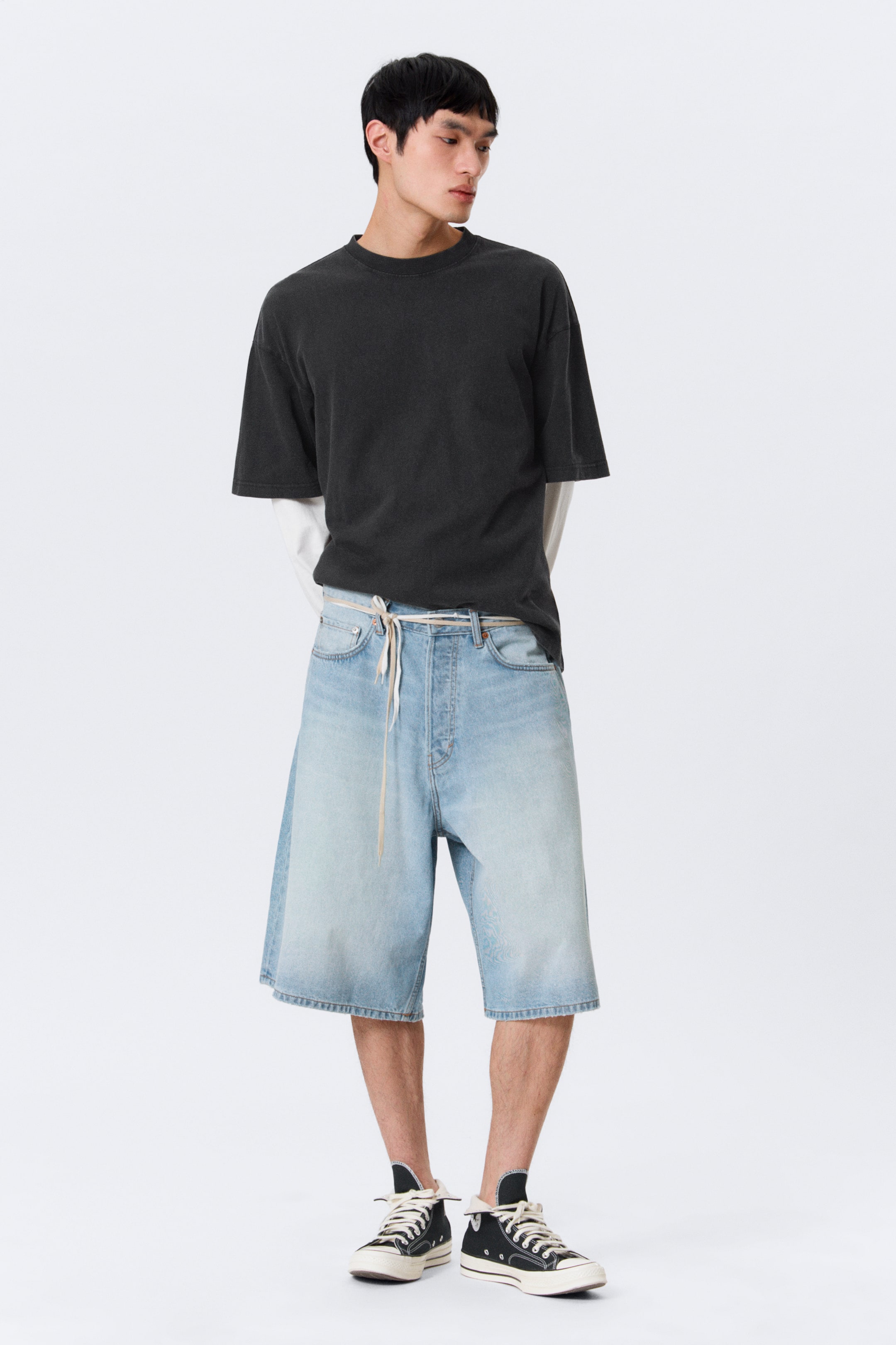 Agrandir l'image: Shorts en denim bleu clair, taille mi-haute, avec une coupe ample et décontractée et des ourlets repliés, portés par le mannequin. Une cordelette de couleur claire est nouée autour de la taille des shorts, qui présentent une braguette à boutons visible et des poches avant ; un t-shirt foncé est partiellement rentré dans la ceinture.