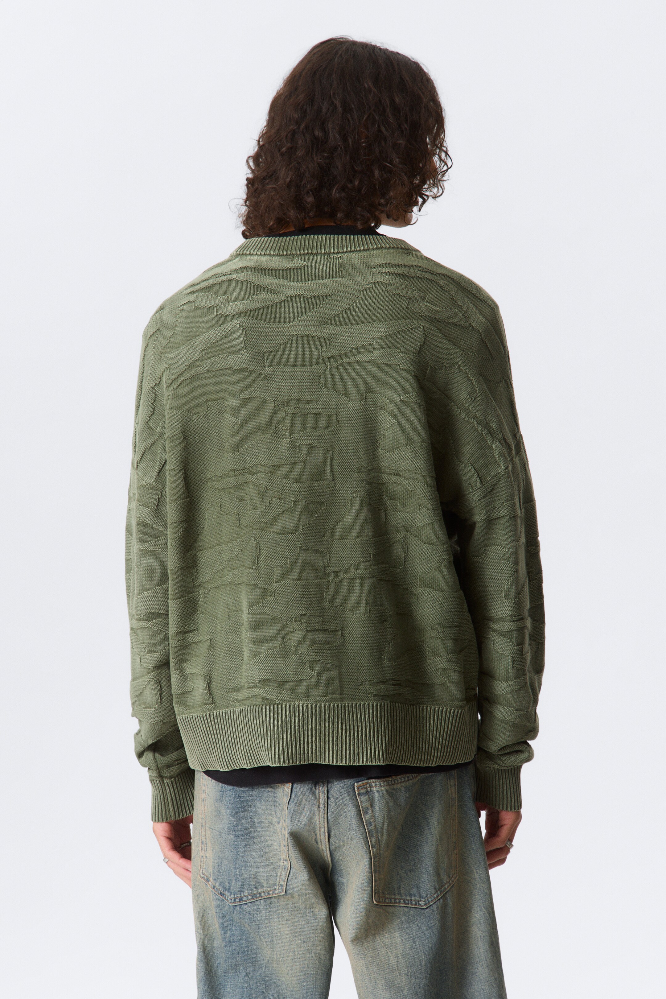 Visa större bild: Den gröna tröjan ses från baksidan och visar ett heltäckande texturerat mönster av oregelbundna, abstrakta former. Den har ribbade muddar och en ribbad kant, med en svart t-shirt synlig som lager under vid halsen och kanten.