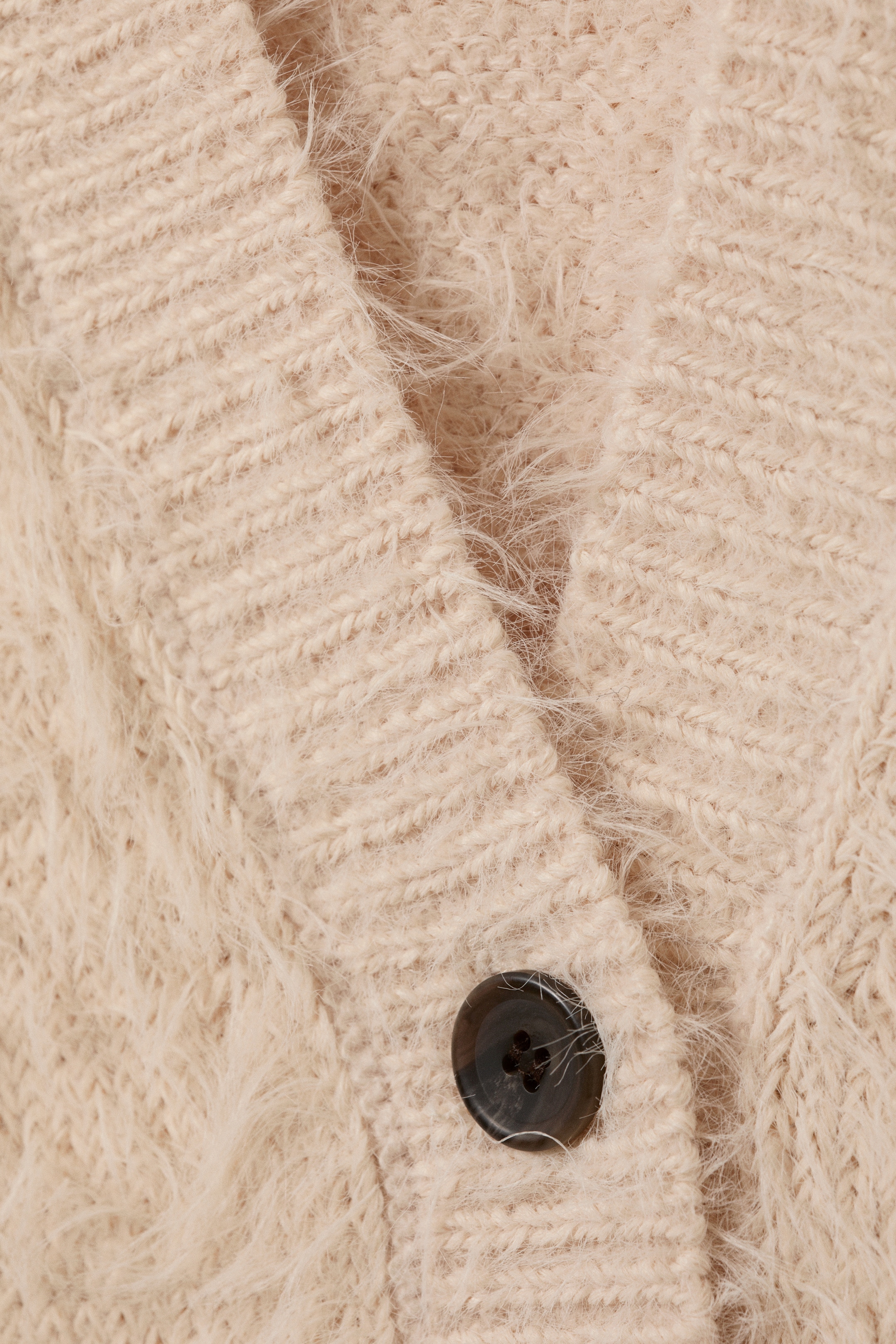 Grotere afbeelding bekijken: Een beige gebreid vest toont een pluizige textuur met een opvallende geribbelde voorbies. Een donkere, viergatsknoop bevestigt de bies, die schuin omhoog loopt om een V-hals opening te vormen.