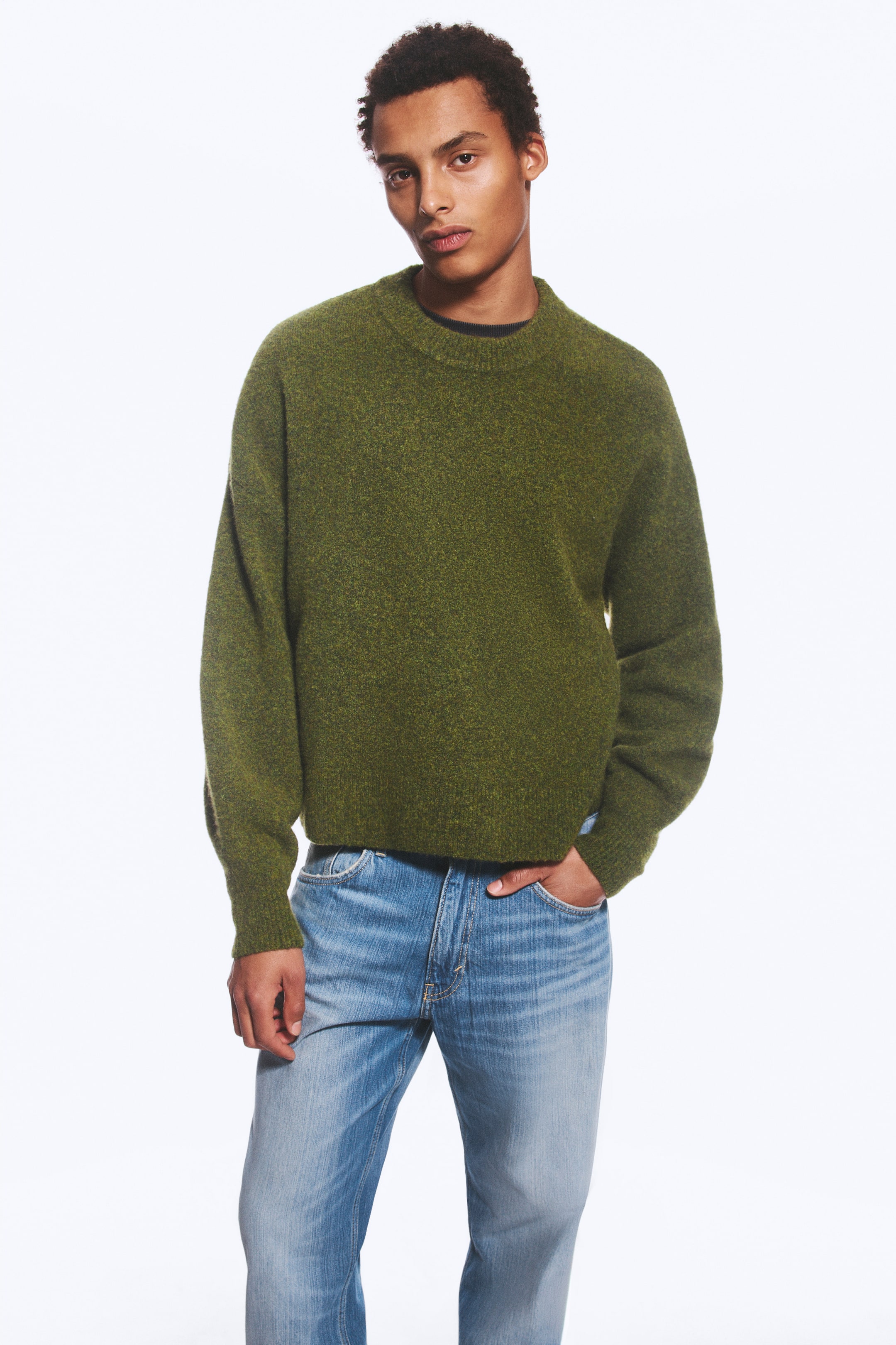 Größeres Bild anzeigen: Ein grün melierter Strickpullover aus Wollmischung mit lockerer Passform, überschnittenen Schultern sowie geripptem Rundhalsausschnitt und Bündchen wird vom Model getragen. Der Pullover wird mit hellblauer Jeans in leichter Waschung kombiniert, und das Model steht mit einer Hand in der vorderen Hosentasche.
