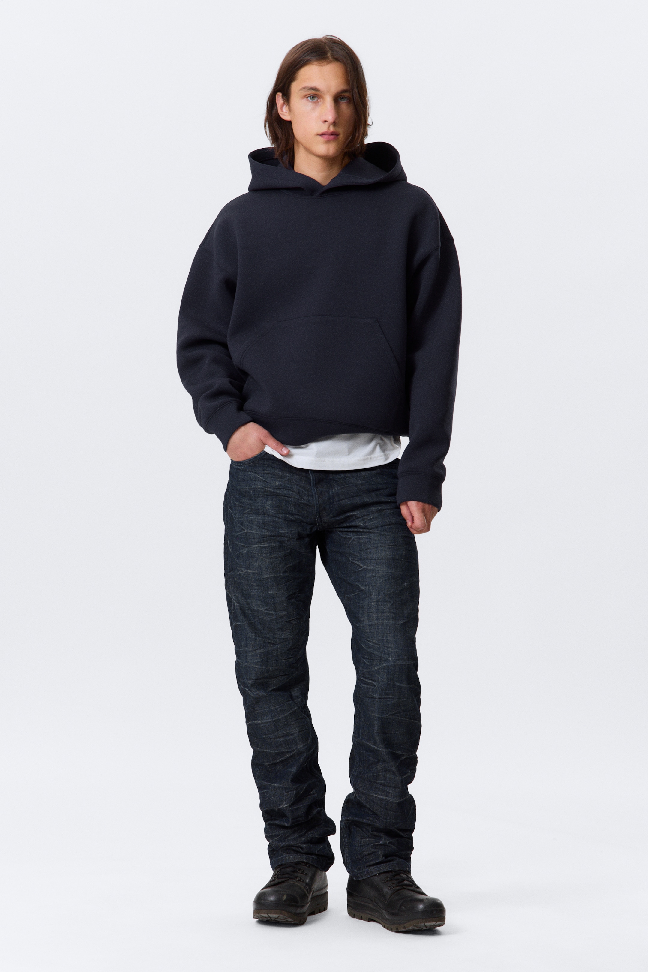 Größeres Bild anzeigen: Ein dunkelblauer übergroßer Hoodie, mit überschnittenen Schultern, einer großen Kapuze und einer auffälligen Kängurutasche, wird von einer Person getragen, die nach vorne schaut, wobei ein weißes T-Shirt unter dem Saum hervorschaut. Dunkel gewaschene Jeans mit einer entspannten Passform und natürlicher Faltenbildung werden ebenfalls präsentiert.
