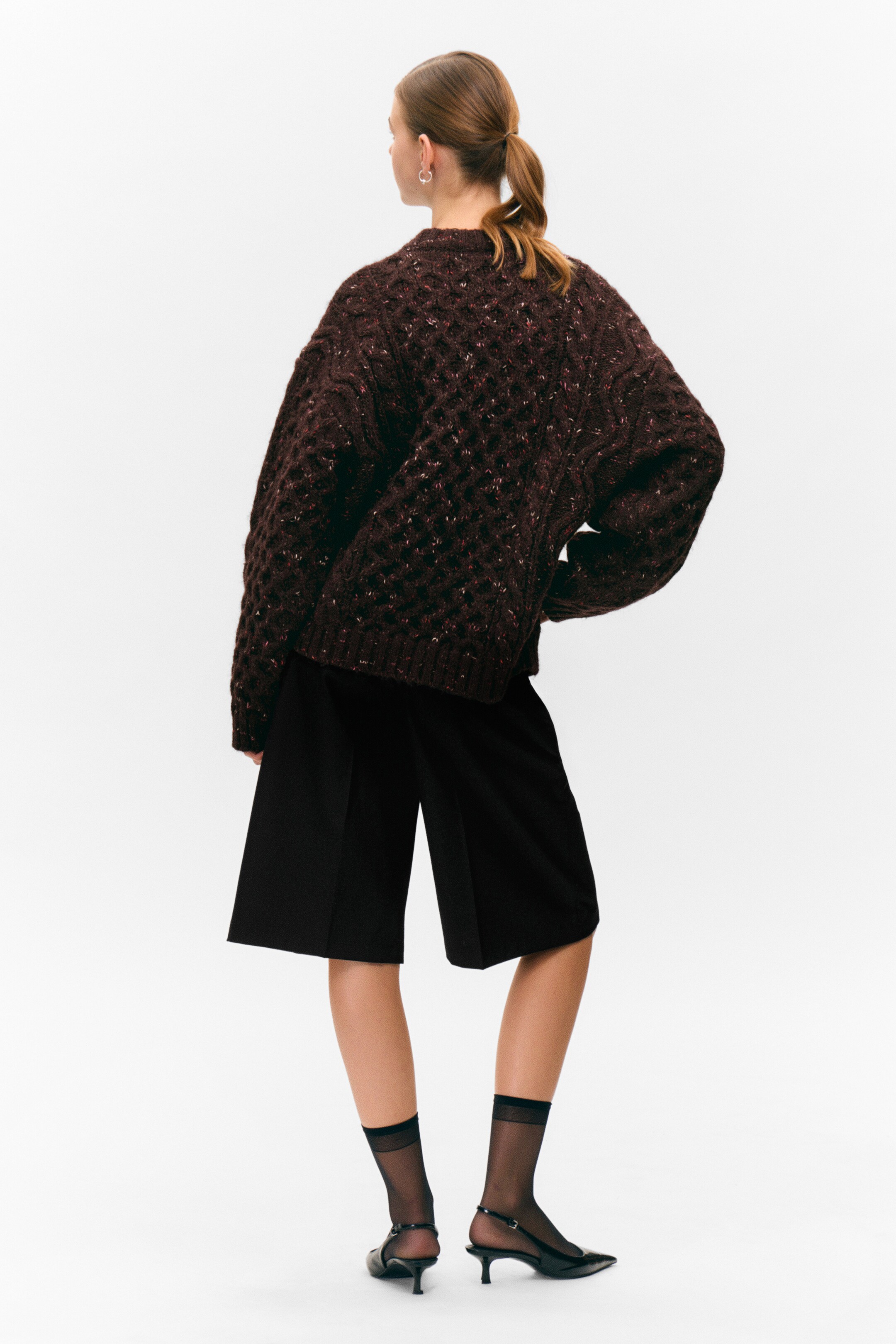 Se større billede: En mørkebrun, tekstureret striktrøje præsenteres fra bagsiden og viser en oversized pasform. Den har et hævet, diamantlignende mønster over hele stoffet med farverige pletter.