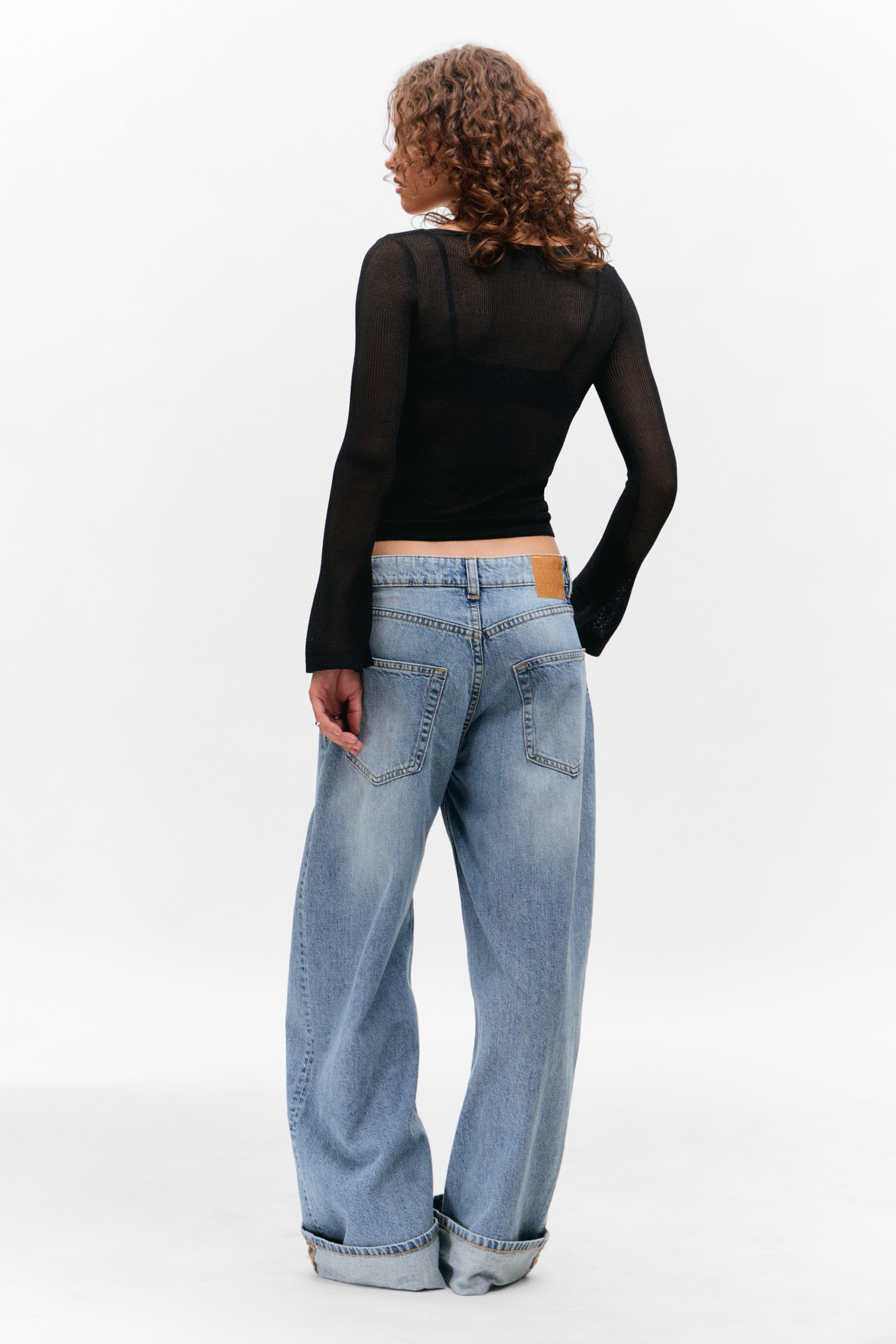 Größeres Bild anzeigen: Ein schwarzes, durchsichtiges, langärmliges Oberteil wird von hinten gezeigt, getragen über einem schwarzen Bralette und mit einem quadratischen Ausschnitt. Hellblaue, weit geschnittene Jeans werden getragen, mit einer tief sitzenden Passform, Gesäßtaschen, einem braunen Aufnäher am Bund und weit aufgerollten Bündchen an den Knöcheln.