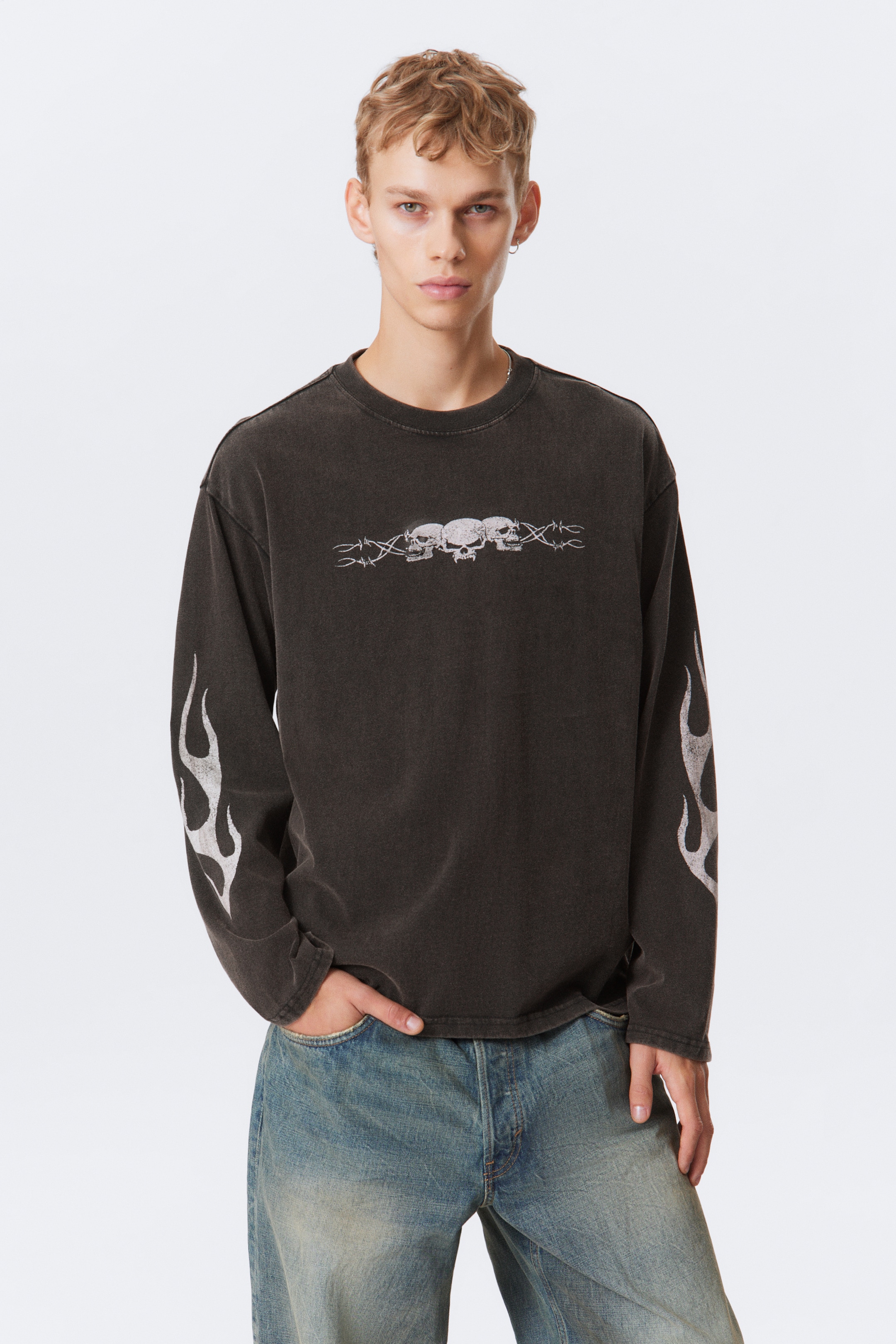Ingrandisci l'immagine: Una camicia nera lavata a maniche lunghe e con una vestibilità ampia è presentata dall'anteriore. Presenta un'immagine grafica di tre teschi danneggiati collegati da filo spinato sul petto, e dei disegni in fiamme bianche appaiono sulla parte esterna di entrambe le maniche.