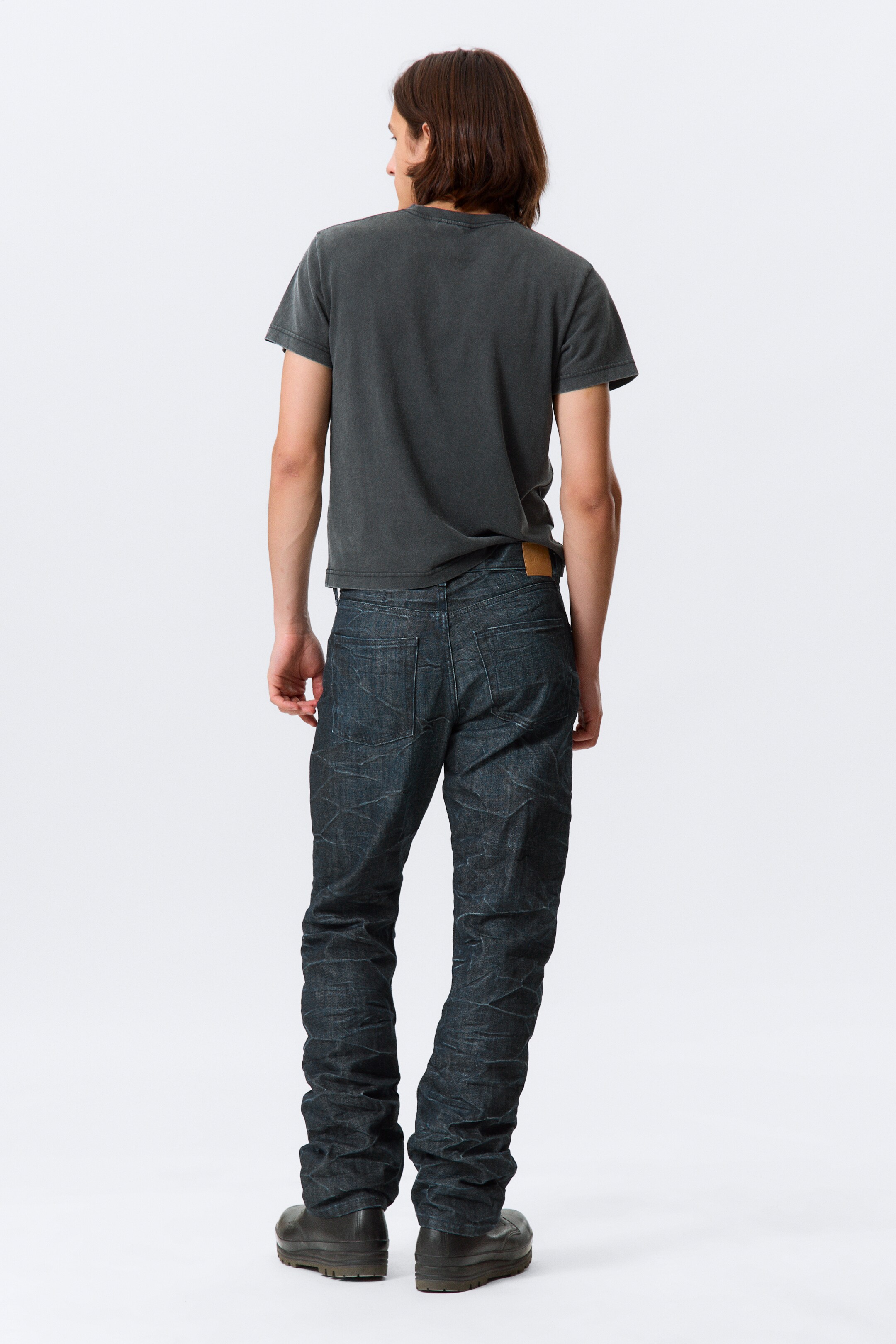 Se større billede: En mørkegrå, vasket t-shirt med rund hals vises fra bagsiden, med korte ærmer. Den er stylet med mørkvaskede jeans, som har en krøllet tekstur, to baglommer og en lysebrun lap på højre side af baglinningen, med benene stablet ved anklerne.