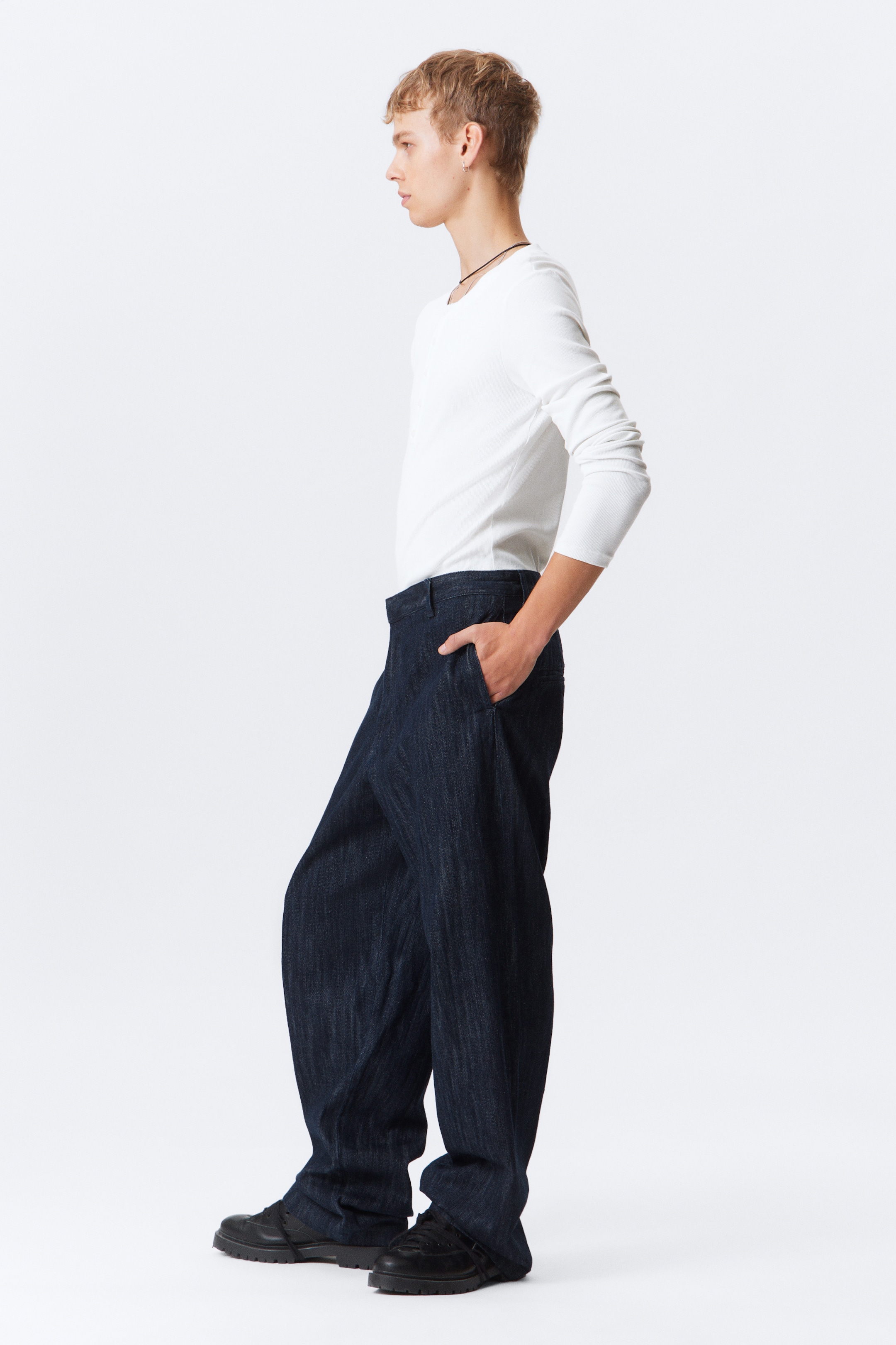 Ver imagen más grande: Los pantalones de mezclilla de lavado oscuro se muestran desde un perfil lateral, con un corte de pierna ancha y un ajuste holgado que se acumula ligeramente sobre el calzado. Los pantalones muestran bolsillos laterales visibles y una cintura sencilla.