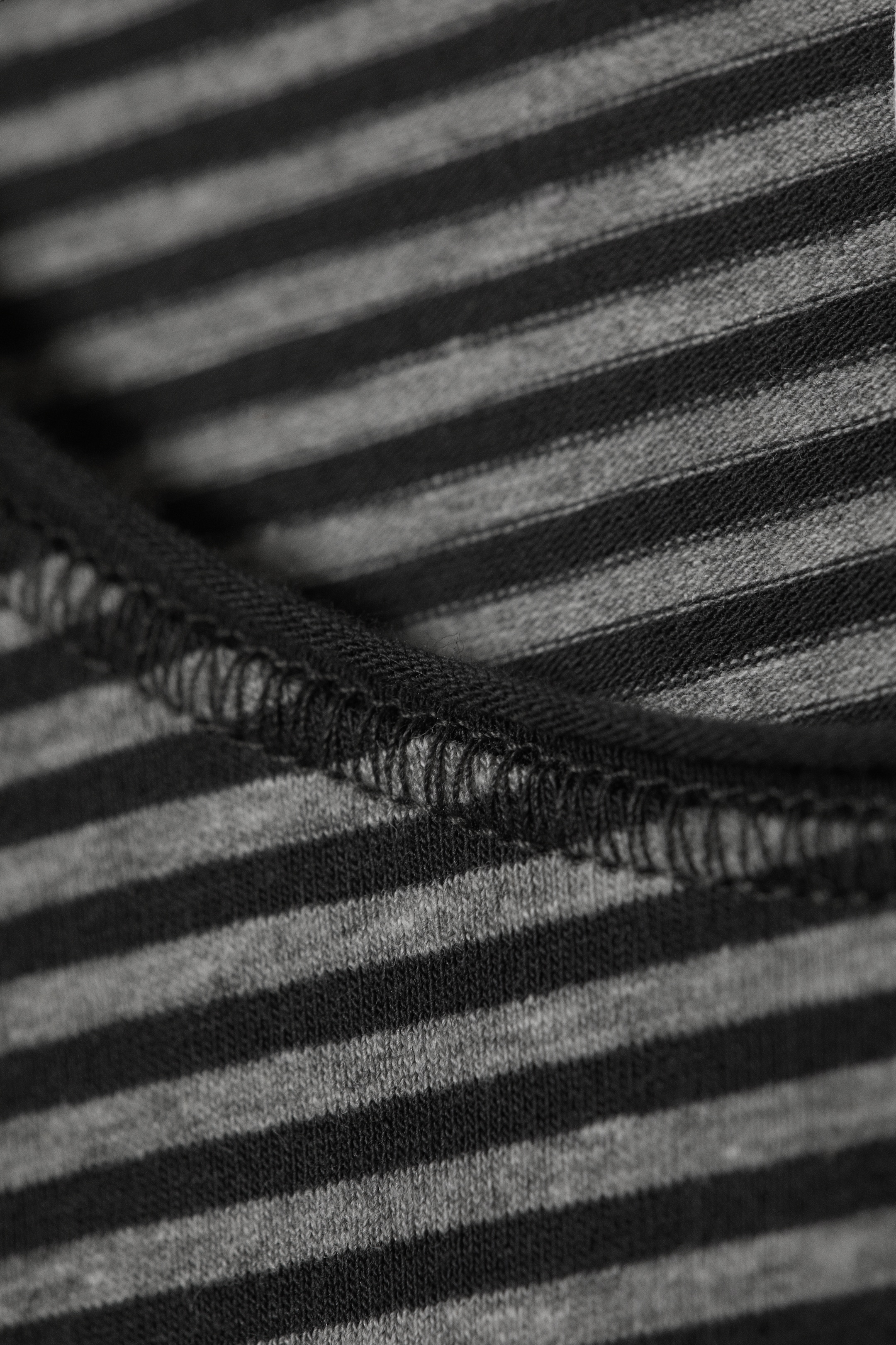 Ingrandisci l'immagine: La maglia a maniche lunghe presenta un primo piano del suo tessuto in jersey, che mostra strisce orizzontali grigio scuro e grigio chiaro alternate. Una cucitura scura e prominente, rifinita a overlock, corre orizzontalmente attraverso il tessuto, mostrando un dettaglio di costruzione.