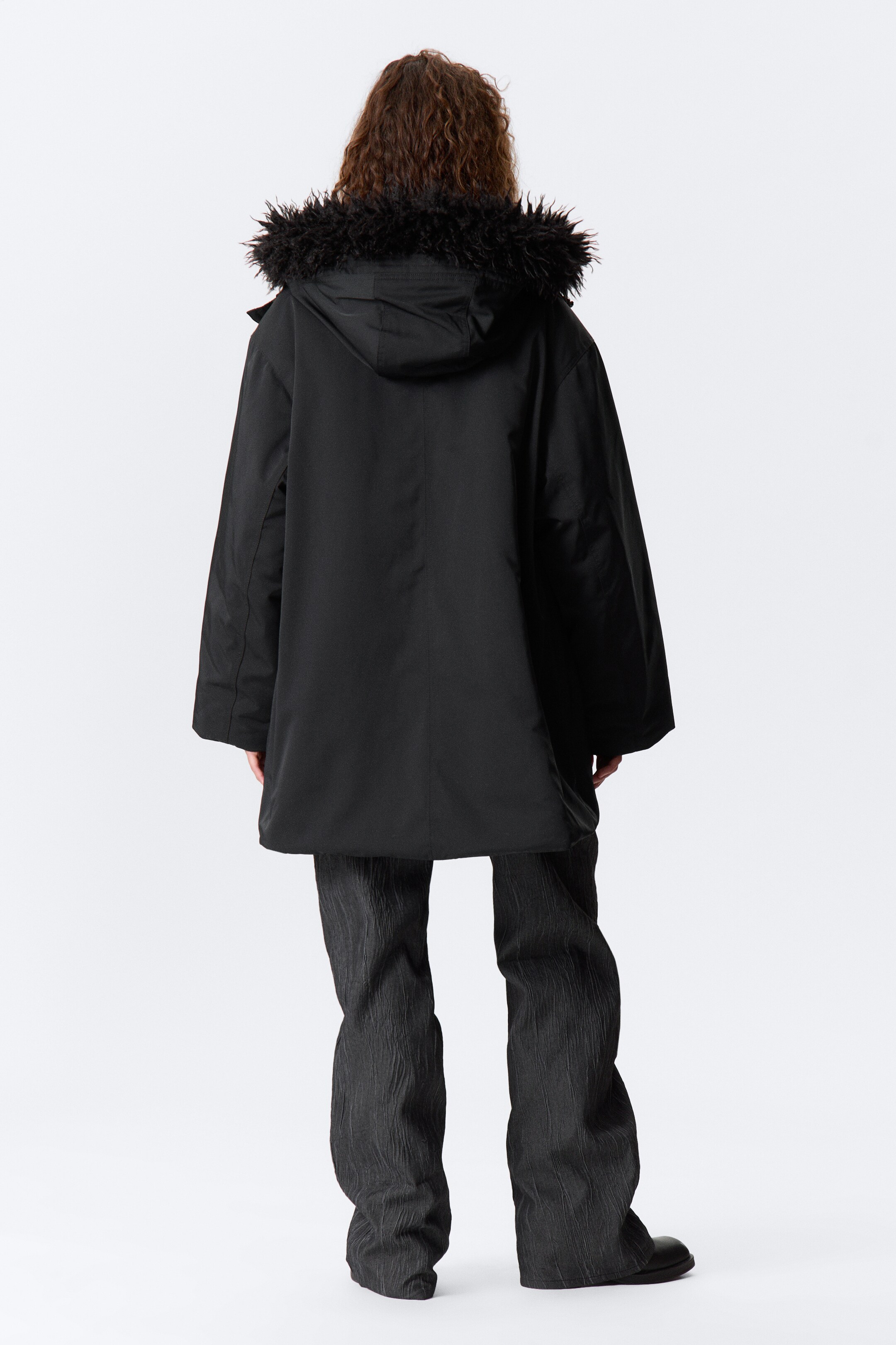 Agrandir l'image: Une parka noire est présentée de dos, avec une capuche ornée d'une garniture en fourrure synthétique noire et une silhouette décontractée de longueur mi-longue. Un pantalon large et texturé de couleur sombre est porté en dessous.