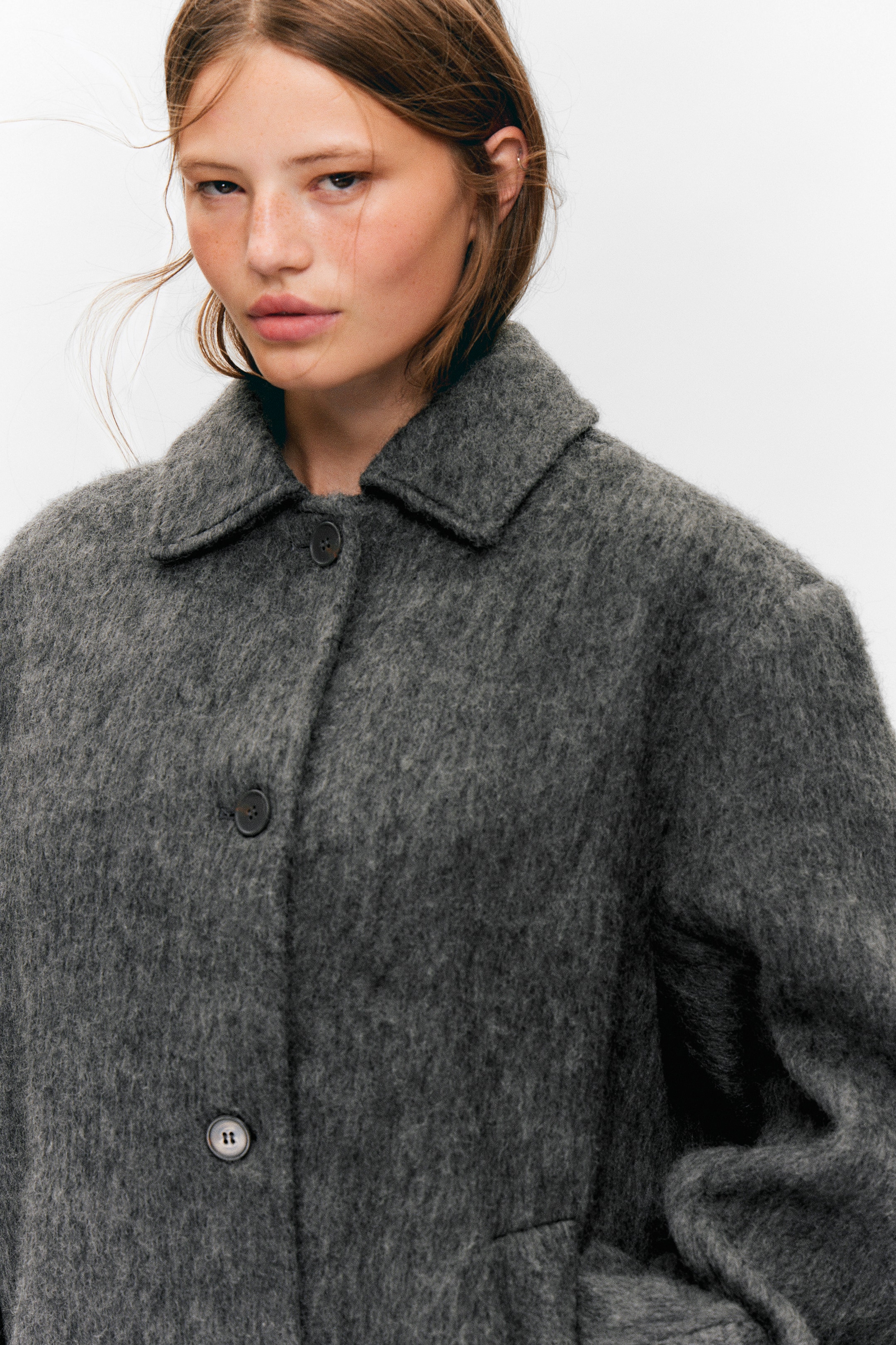Ingrandisci l'immagine: Un cappotto grigio dalla texture irsuta è presentato da un angolo leggermente frontale-sinistro, caratterizzato da un classico colletto a punta e una chiusura frontale con due bottoni scuri. Una sottile cucitura orizzontale è visibile sulla parte anteriore inferiore, indicando una tasca.