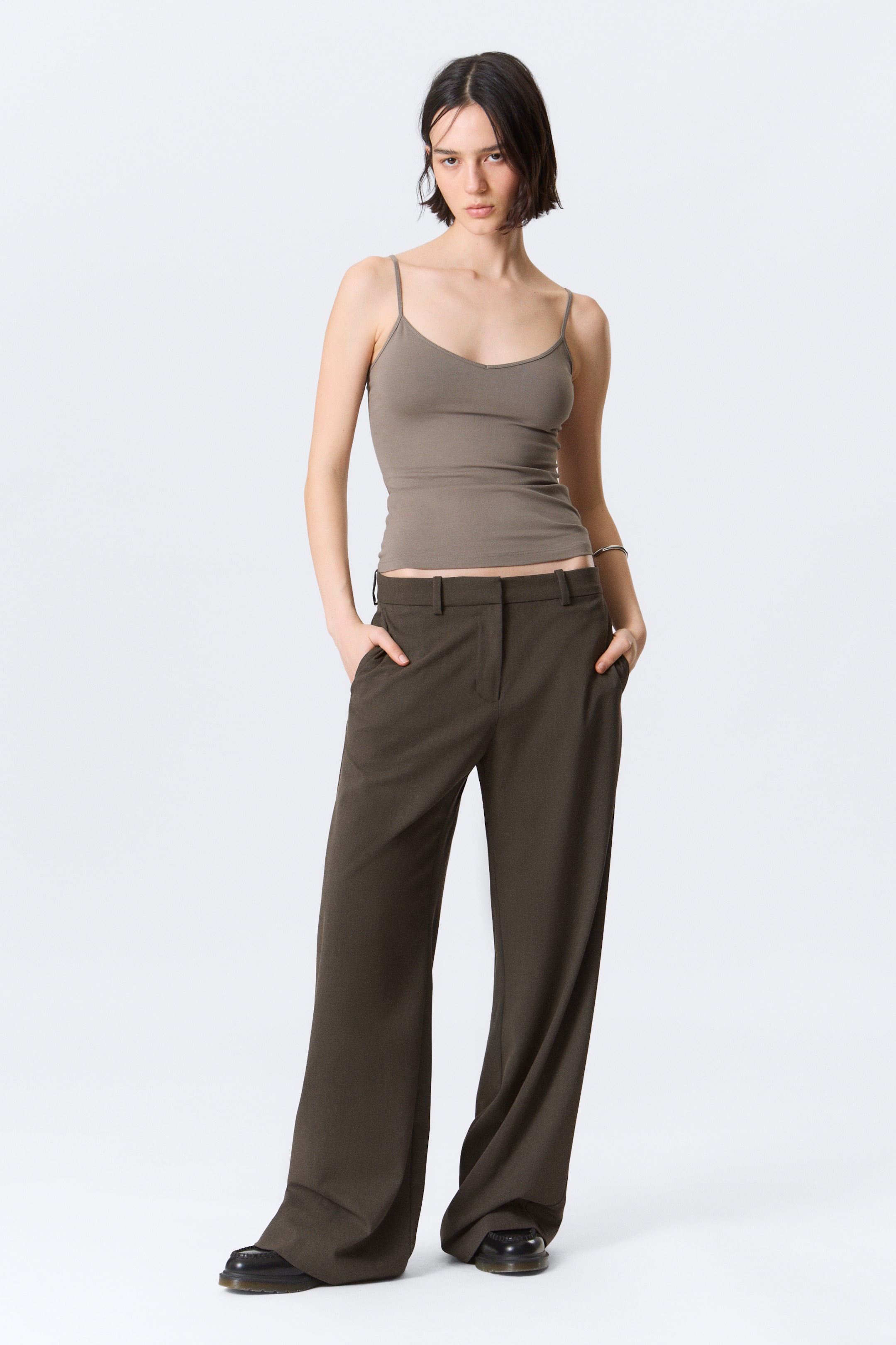 Zobraziť väčší obrázok: "Priliehavý taupe top s tenkými ramienkami a výstrihom do V je nosený, jeho lem dosahuje pás tmavých olivovo hnedých nohavíc so širokými nohavicami. Nohavice sú zobrazené z predného pohľadu, majú zips a zapínanie na gombík, pútka na opasok a bočné vrecká."