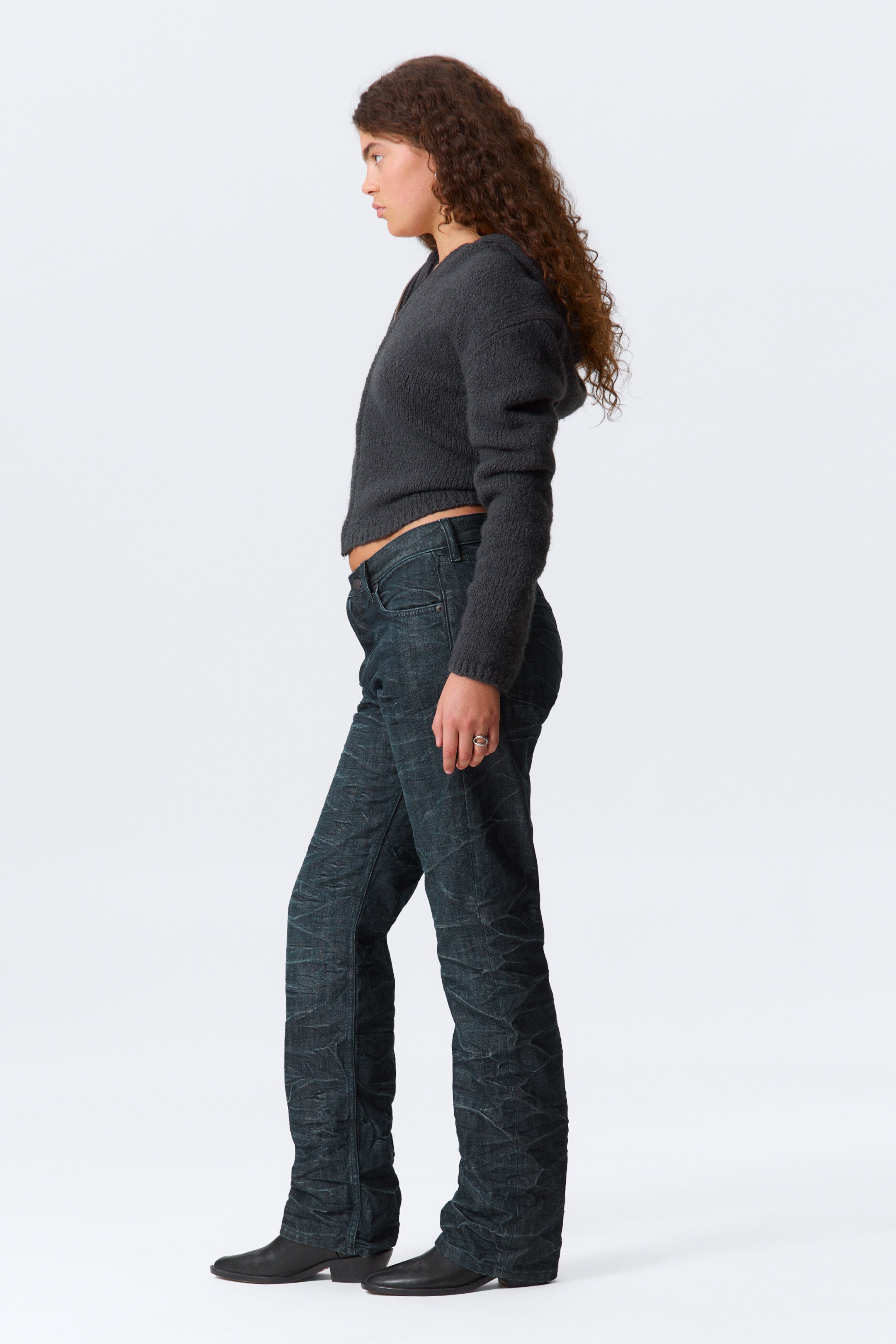 Se større billede: En mørkegrå, beskåret, fuzzy hættetrøje bliver vist i sidevisning, med lange ærmer og en synlig hætte. Mørke vaskede jeans bæres, der viser en krøllet tekstur og en lige benpasform, der sidder lavt på taljen.