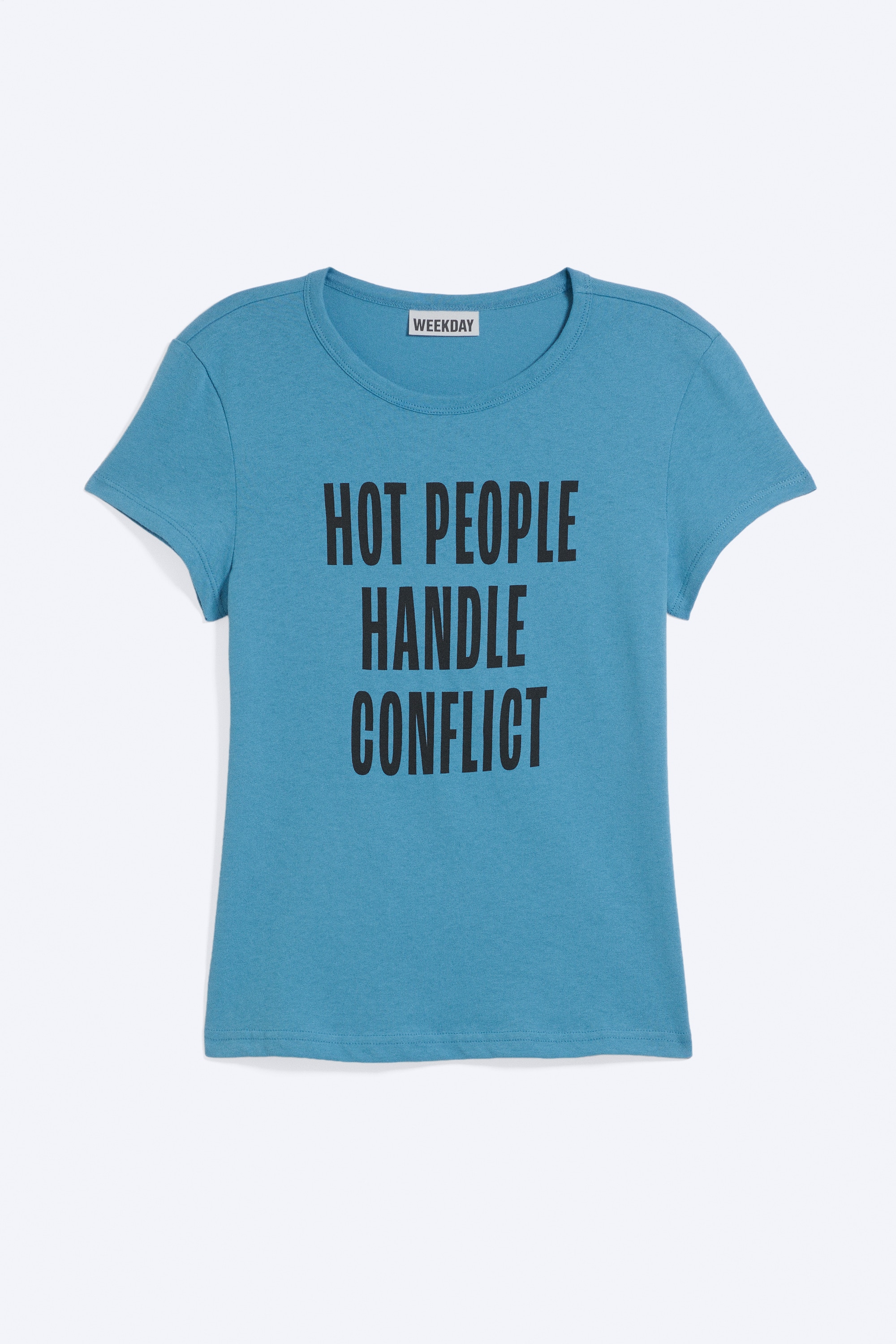 La modelo lleva una camiseta azul de manga corta con cuello redondo, bajo recto y el texto impreso "HOT PEOPLE HANDLE CONFLICT". La camiseta se combina con pantalones negros tobilleros.