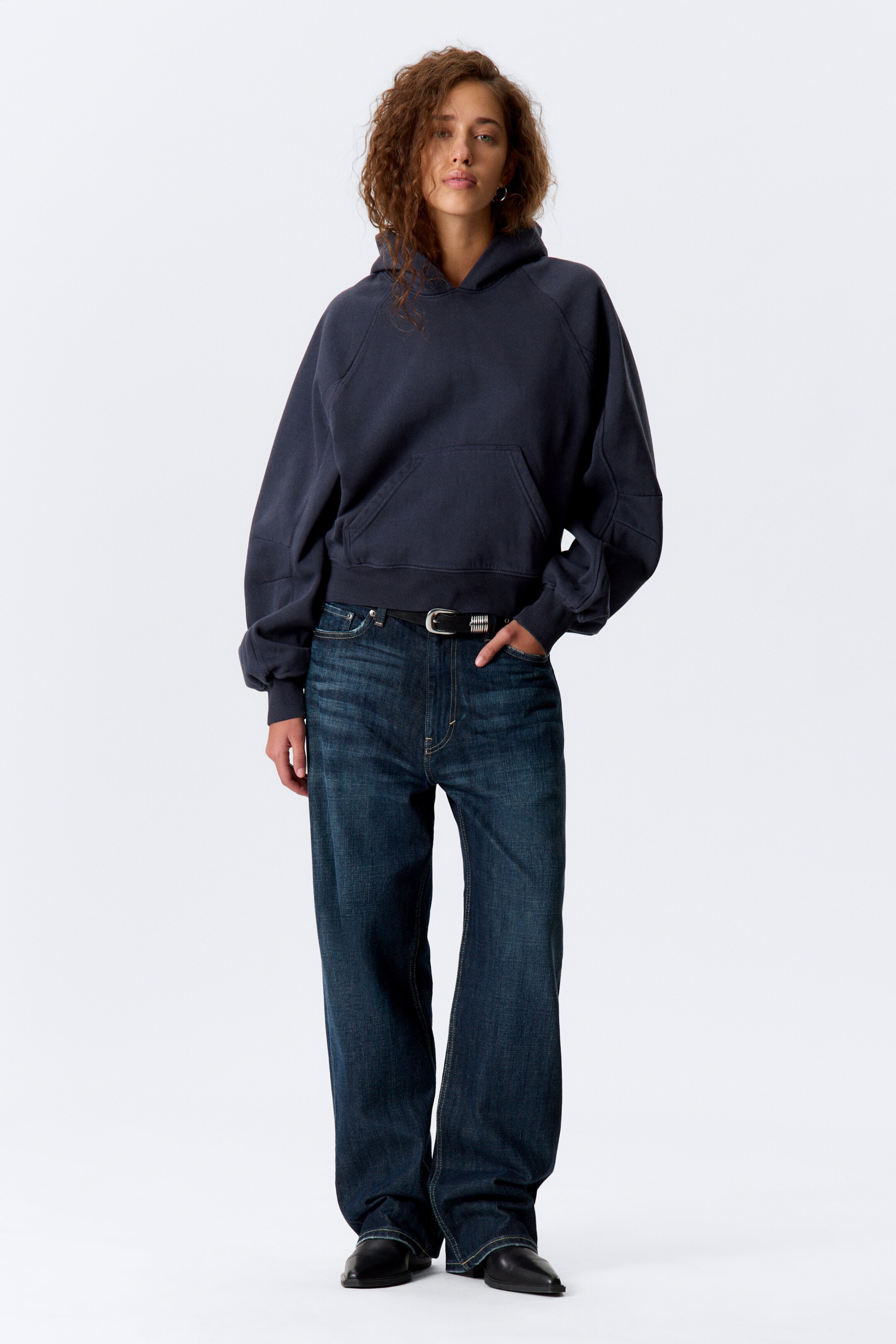 Größeres Bild ansehen: Ein dunkelblauer, übergroßer Hoodie wird von vorne präsentiert, mit Raglanärmeln mit sichtbaren Nahtdetails und gerippten Bündchen. Er zeigt auch eine Kängurutasche vorne und einen gerippten, gekürzten Saum, der über dunkel gewaschene Jeans geschichtet ist.