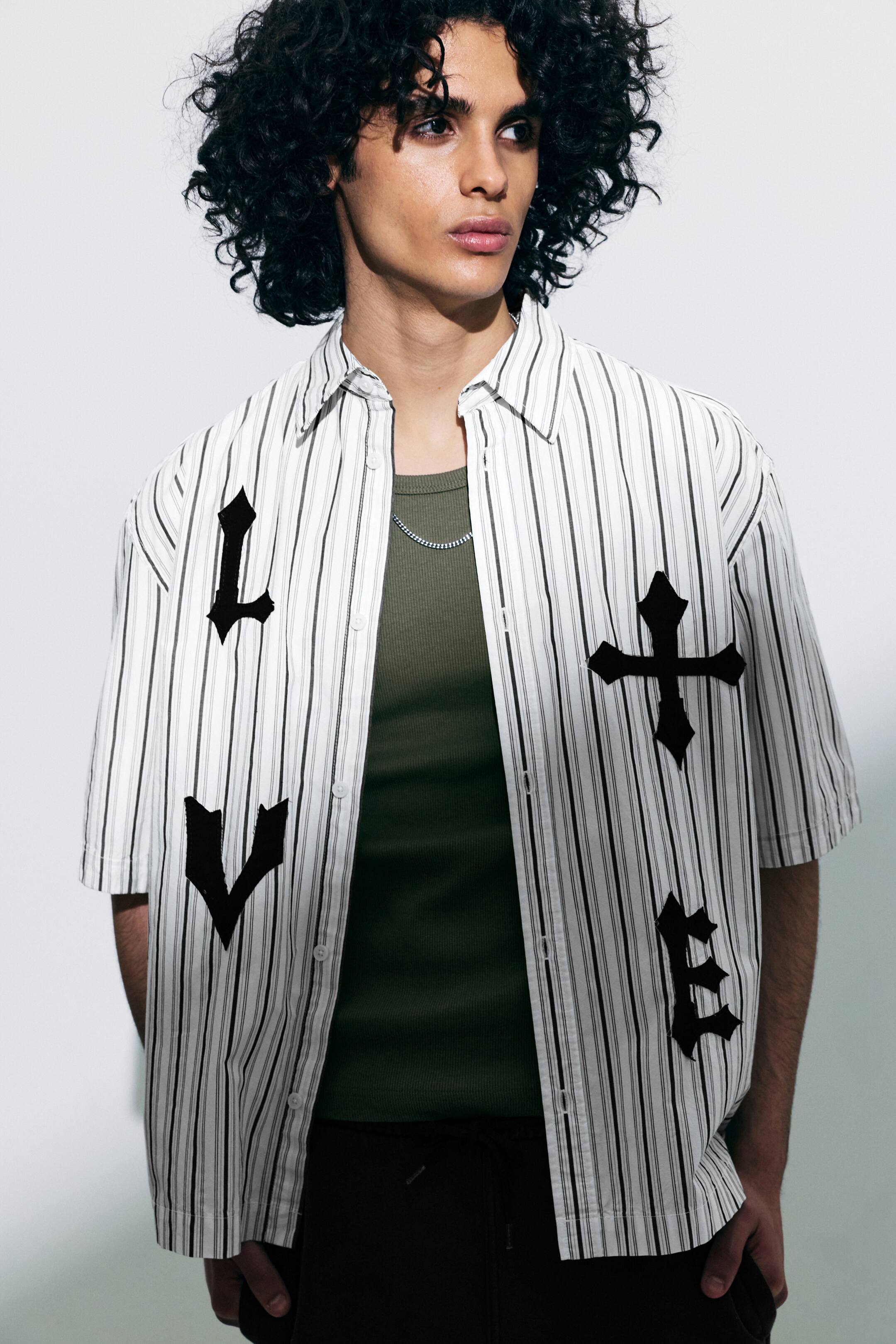 Visualizza l'immagine ingrandita: Una camicia bianca a maniche corte con bottoni, caratterizzata da sottili righe verticali scure, è indossata aperta sopra un top a costine verde scuro, rivelando una catena d'argento. Sul davanti della camicia compaiono lettere in stile gotico nero "L", "V" e "E" insieme a un motivo a croce.