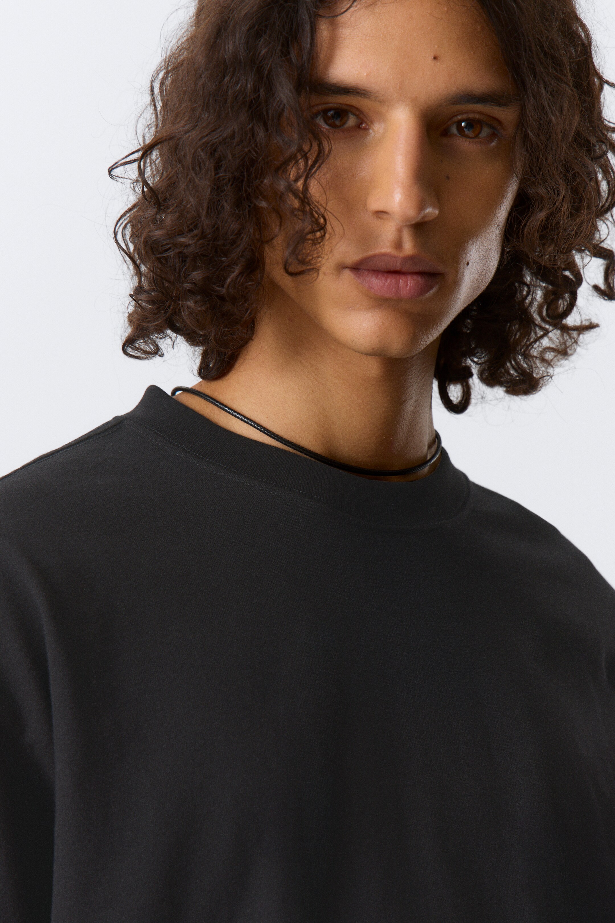 Grotere afbeelding bekijken: Een zwart trui met ronde hals wordt gedragen, iets naar voren gekanteld, en toont zijn gladde, matte stof en geribbelde kraag. Een dun zwart koordketting rust op de halslijn van het shirt.
